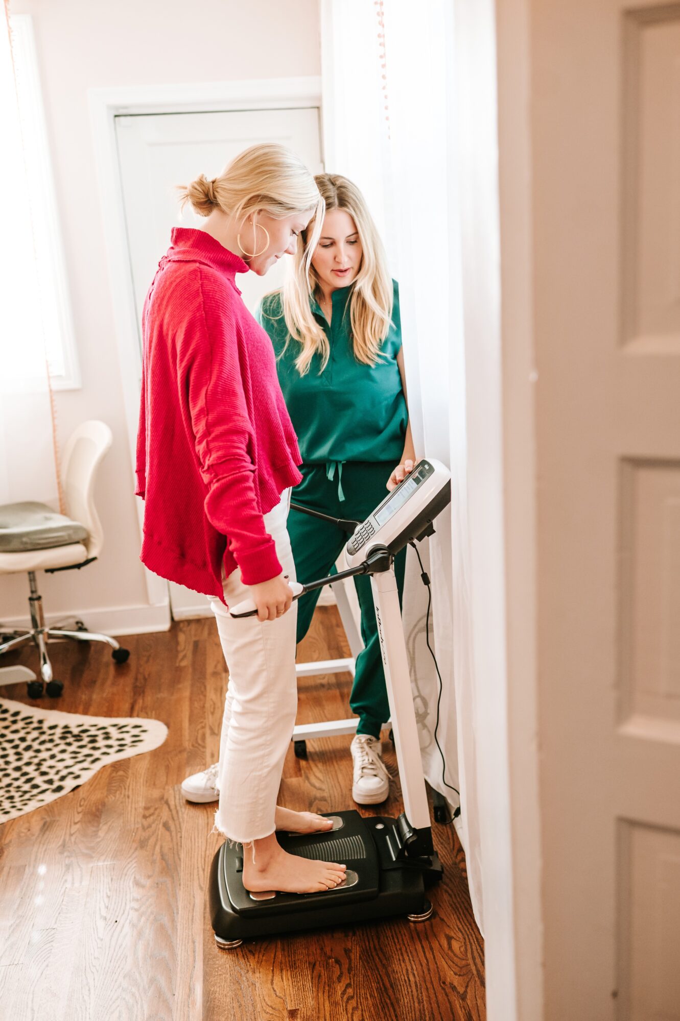 Two women stand on a scale in a room with wooden floor, one in a red jacket and the other in green, looking at the scale display.