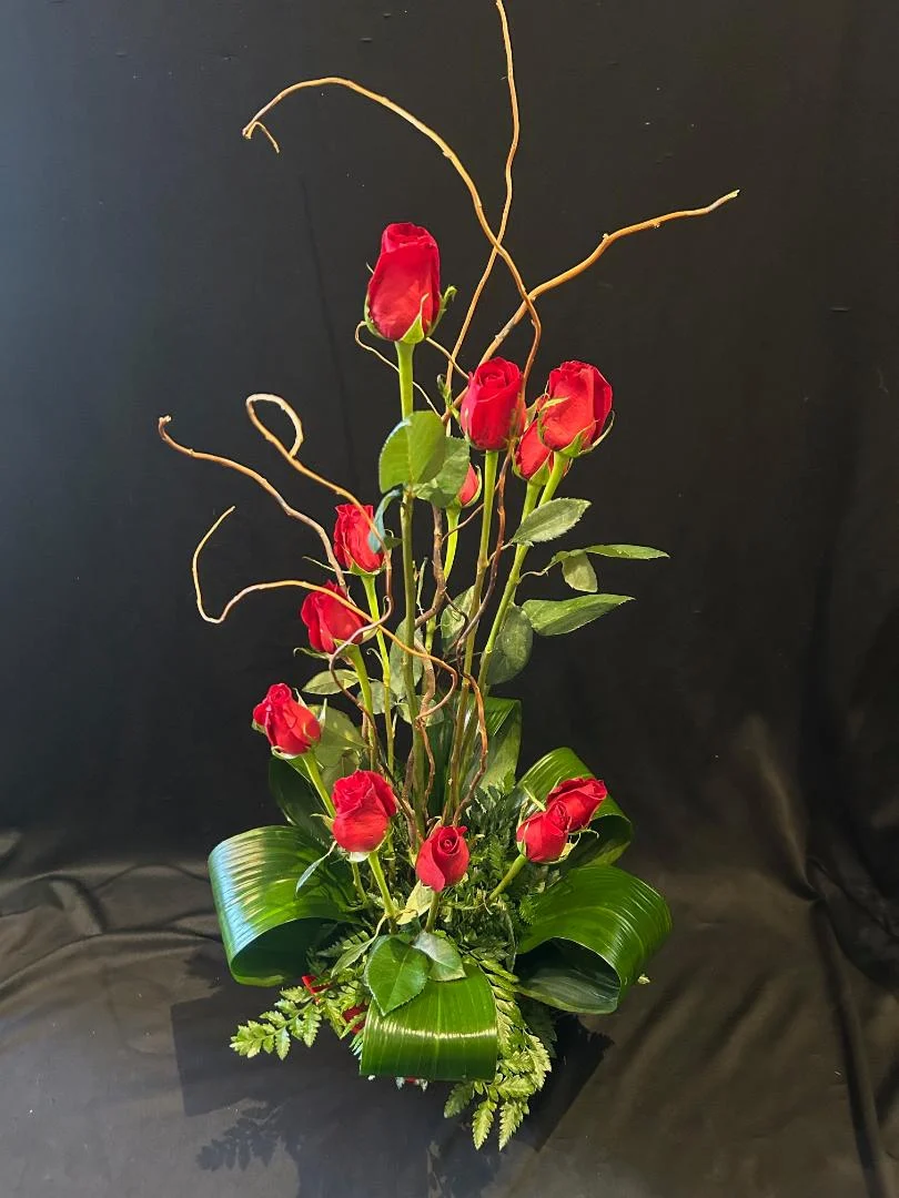 Flower arrangement with red roses, green leaves, and curly twigs in a green container against a dark background.