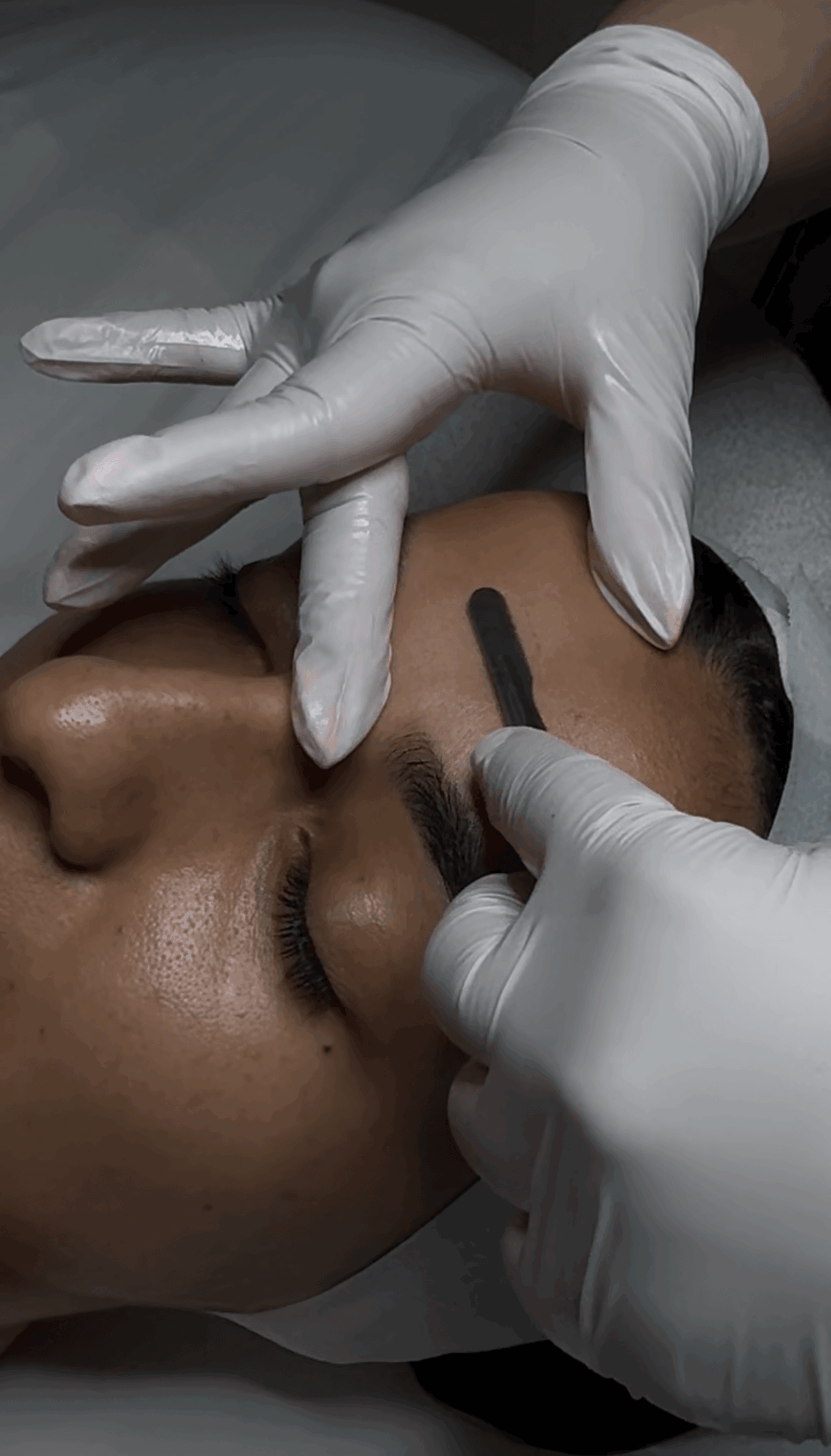 Person receiving eyebrow tattooing with a technician using a tool, wearing gloves, close-up of face and hands, dark background.