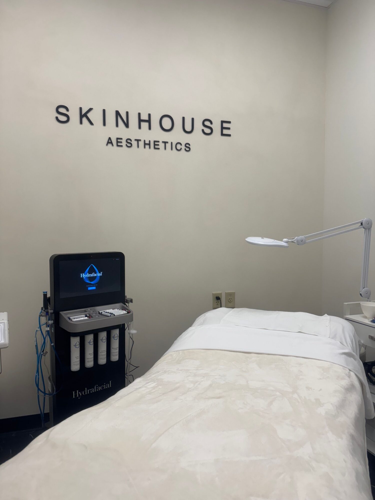 A medical treatment room with a bed, a monitor, and a wall with the words 'SKINHOUSE AESTHETICS'.