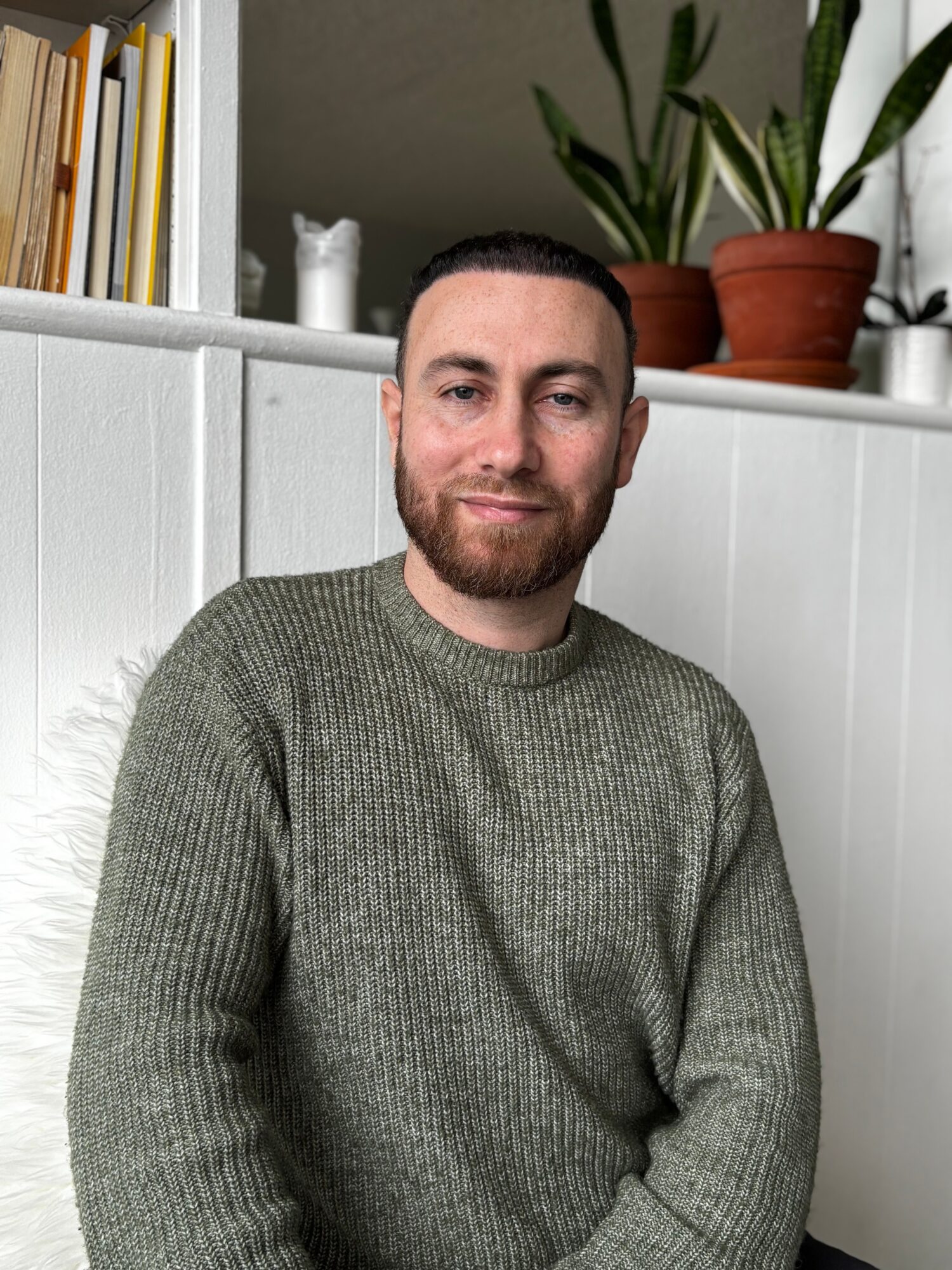 Man with short dark hair and beard, wearing a green sweater, sitting indoors with plants and books behind him.