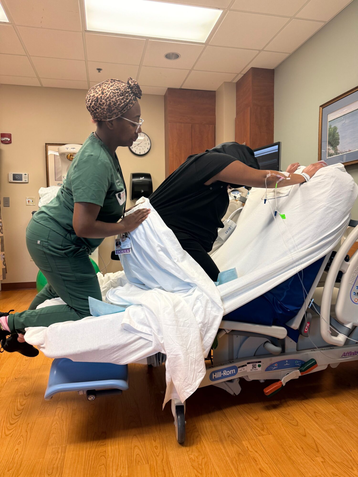 Person lying in hospital bed with medical staff attending, hospital room with wooden floor and medical equipment visible.