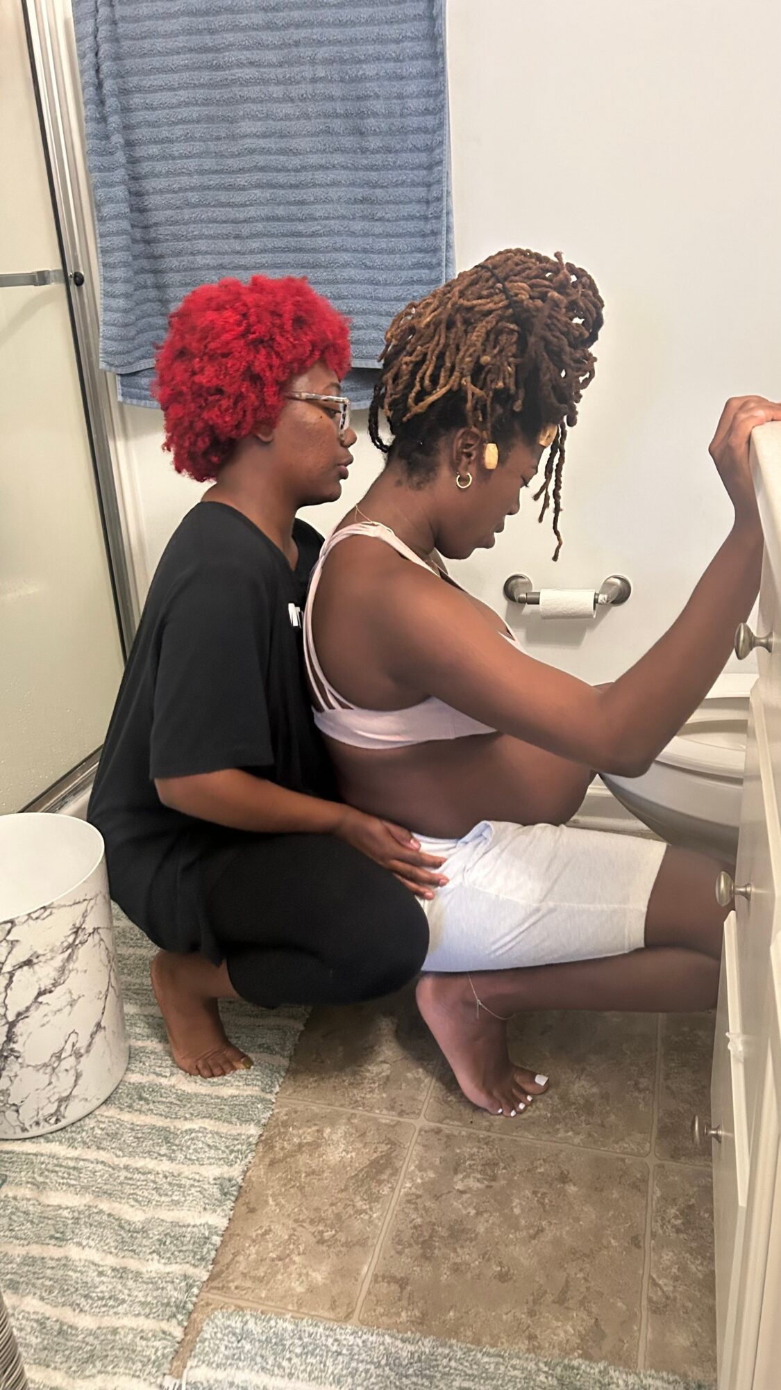 Two women with natural hairstyles sitting on a bathroom floor, one in black clothing and the other in a light top, facing a mirror.
