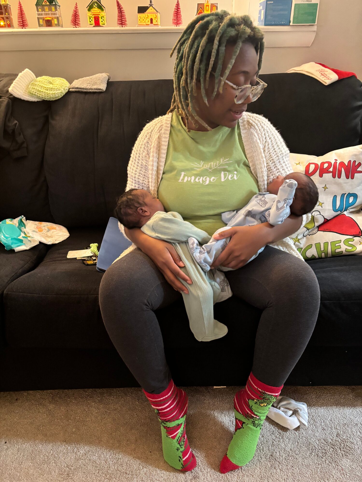 Person sitting on a black couch holding a baby, with colorful socks and a green shirt, in a room with bookshelves.