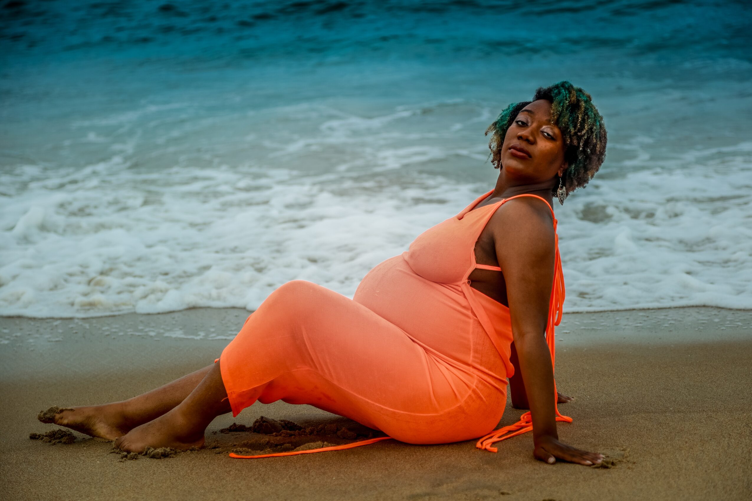 Pregnant woman in orange dress sitting on beach sand near ocean waves, looking at camera, with short curly hair.