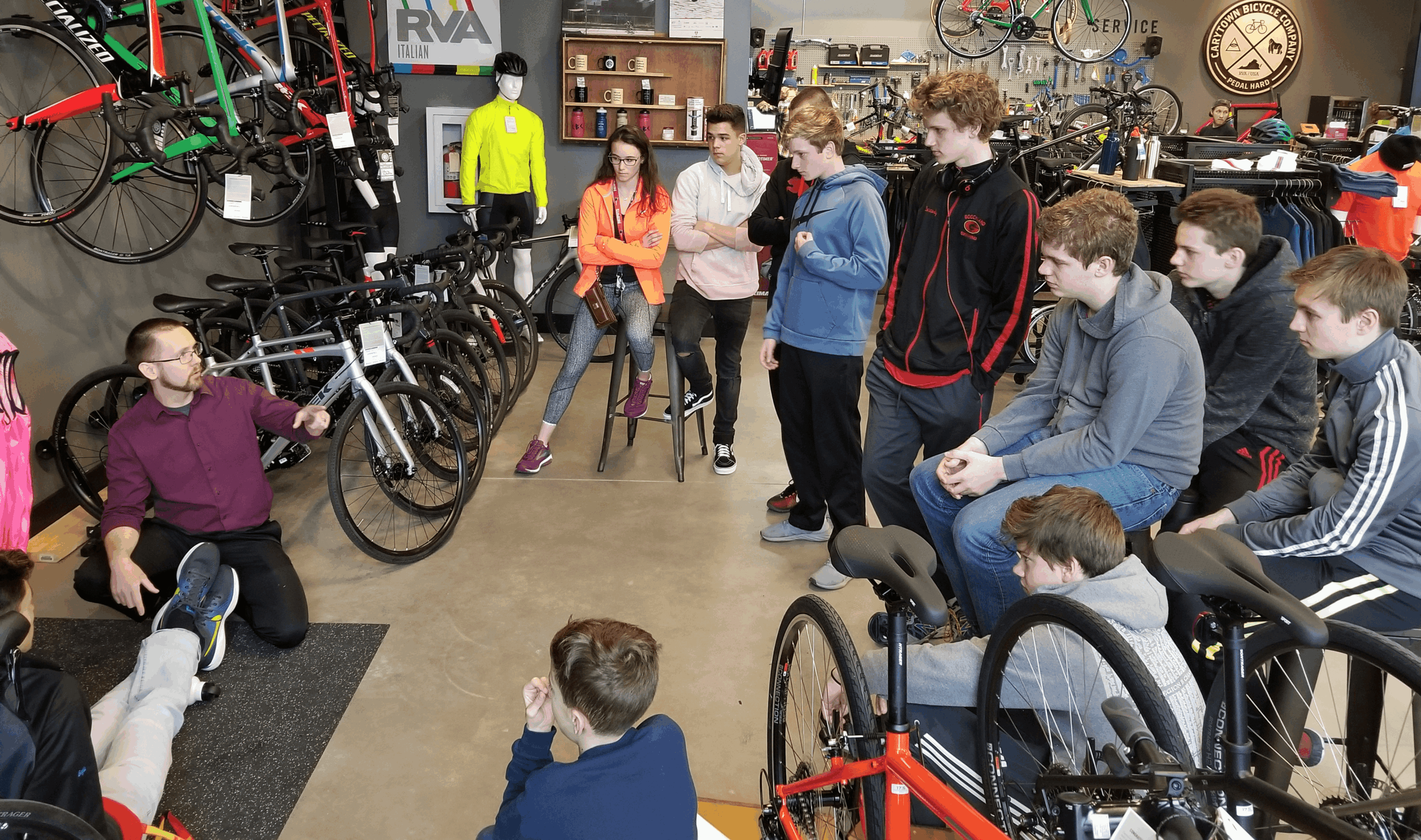 Group of people listening to a woman speaking in a bicycle shop with bikes on the wall and tools displayed.