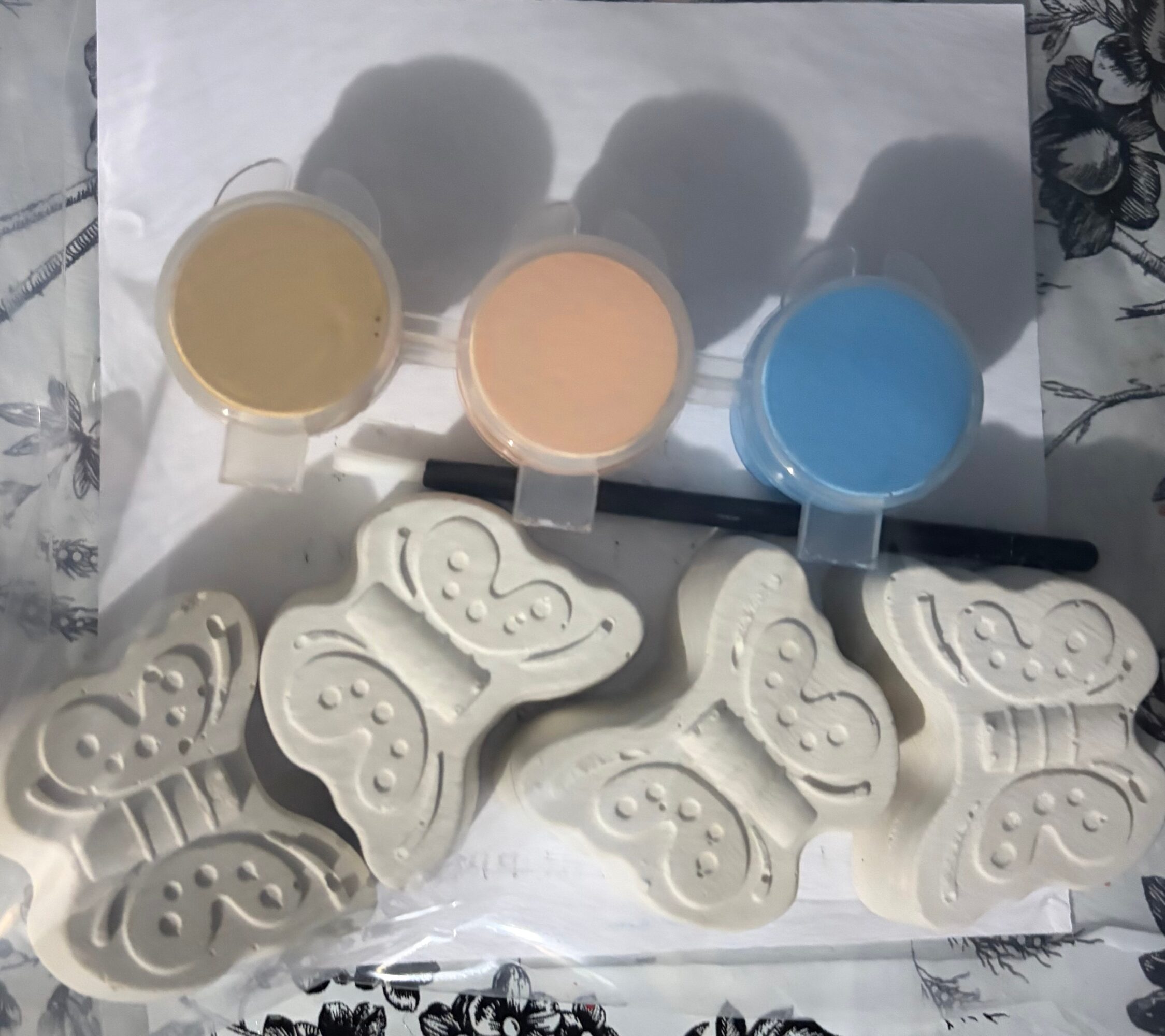 Three small containers of colored paint—yellow, peach, and blue—are above butterfly-shaped soap molds on a surface with floral pattern.
