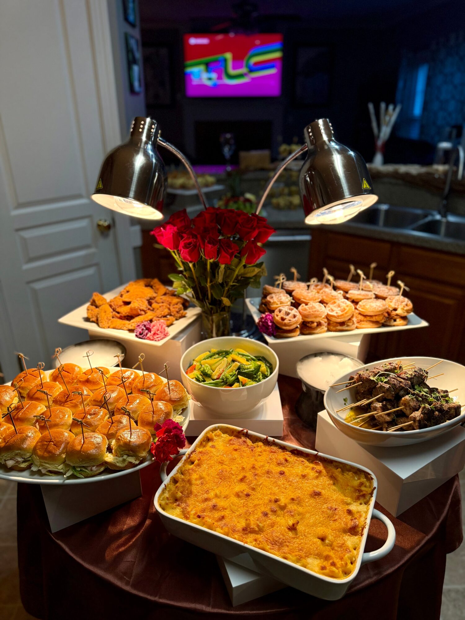 Table with red roses, cupcakes, baked dish, salad, and other food, illuminated by two lamps, in a kitchen setting.