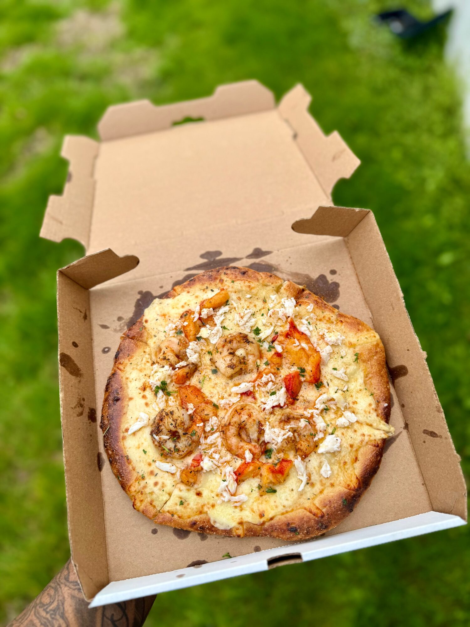 Pizza in an open cardboard box with a green outdoor background.