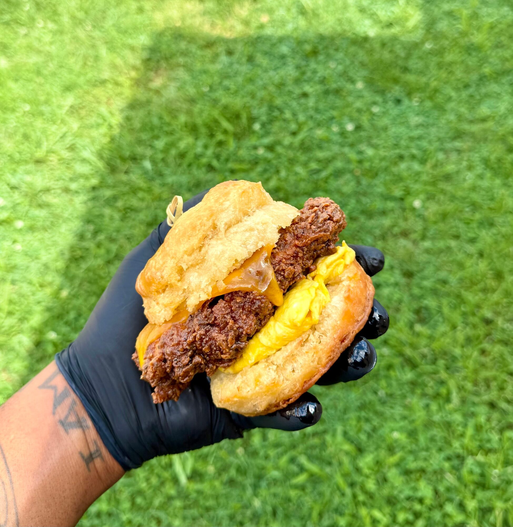 Hand holding a burger with fried chicken, beef patty, cheese, and bun against green grass background.