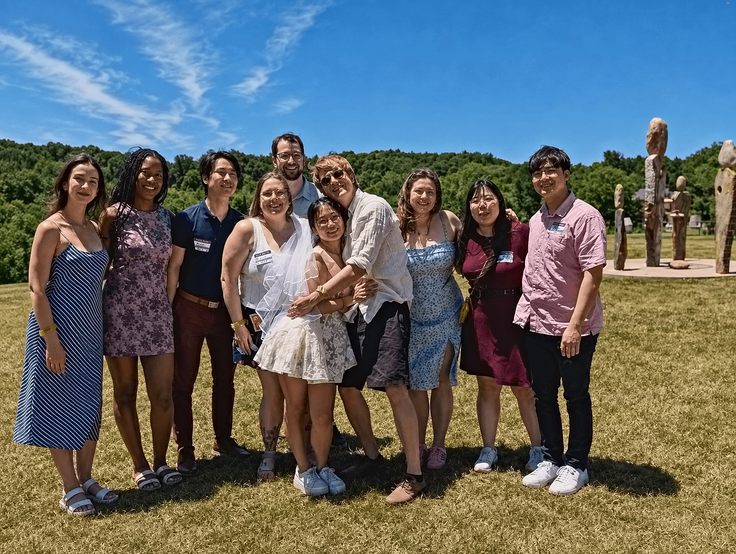 Group of ten people standing outdoors on grass with trees and sculptures in the background under a blue sky.