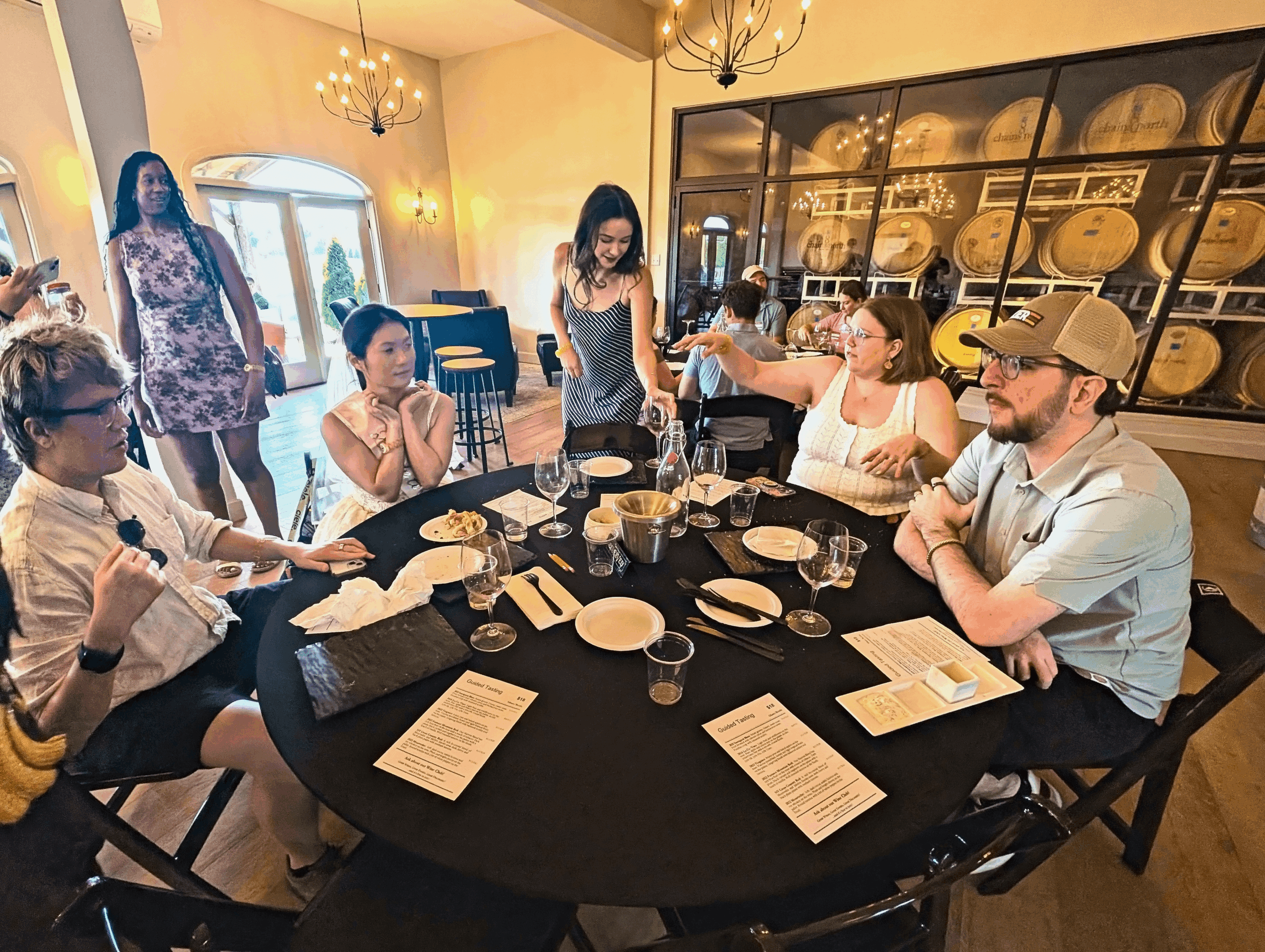 Group of people seated around a table in a room with shelves of dishes, some standing and some sitting, engaged in conversation.