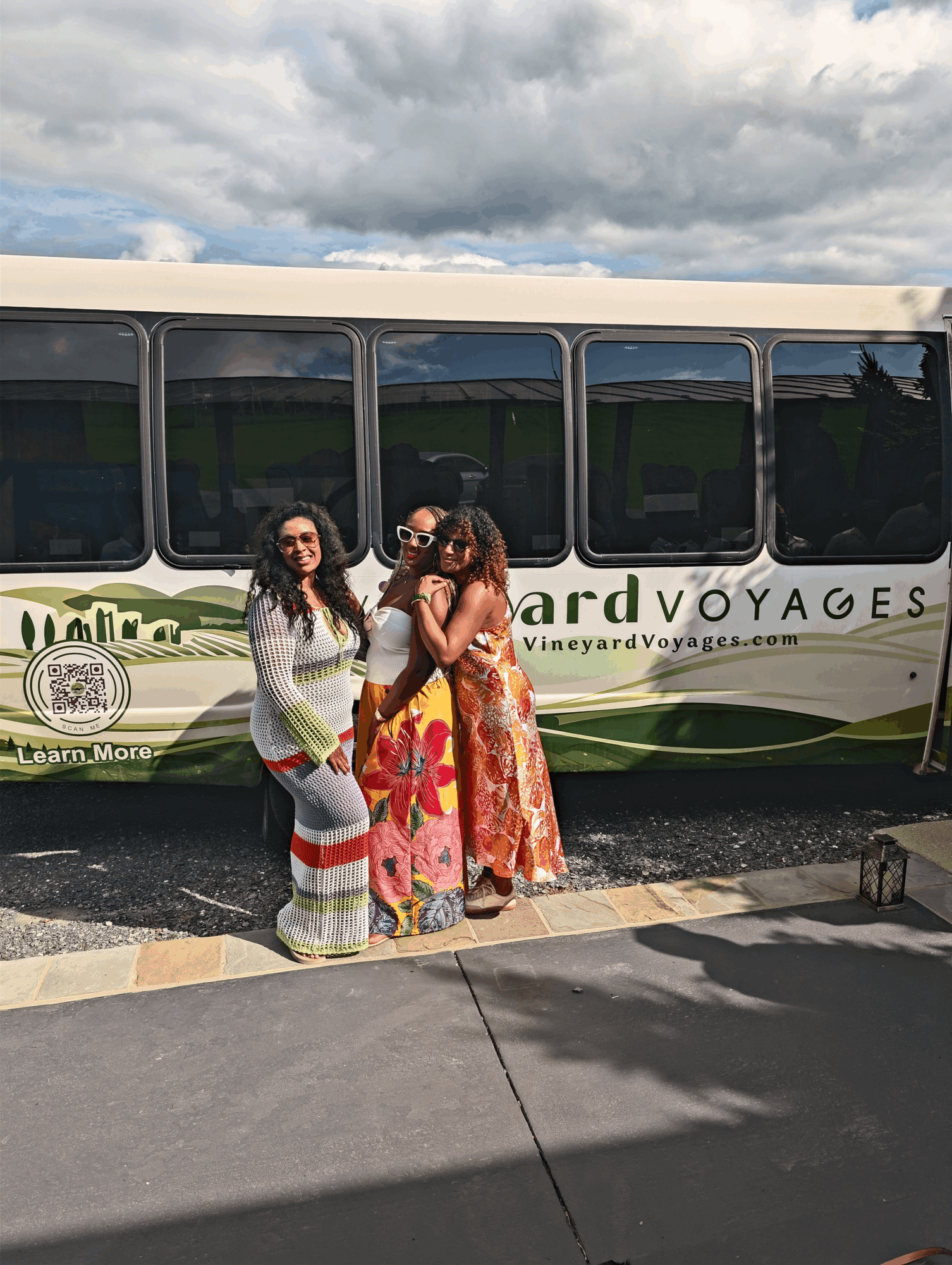 Three women stand in front of a bus with a cloudy sky above, wearing colorful dresses and sunglasses.