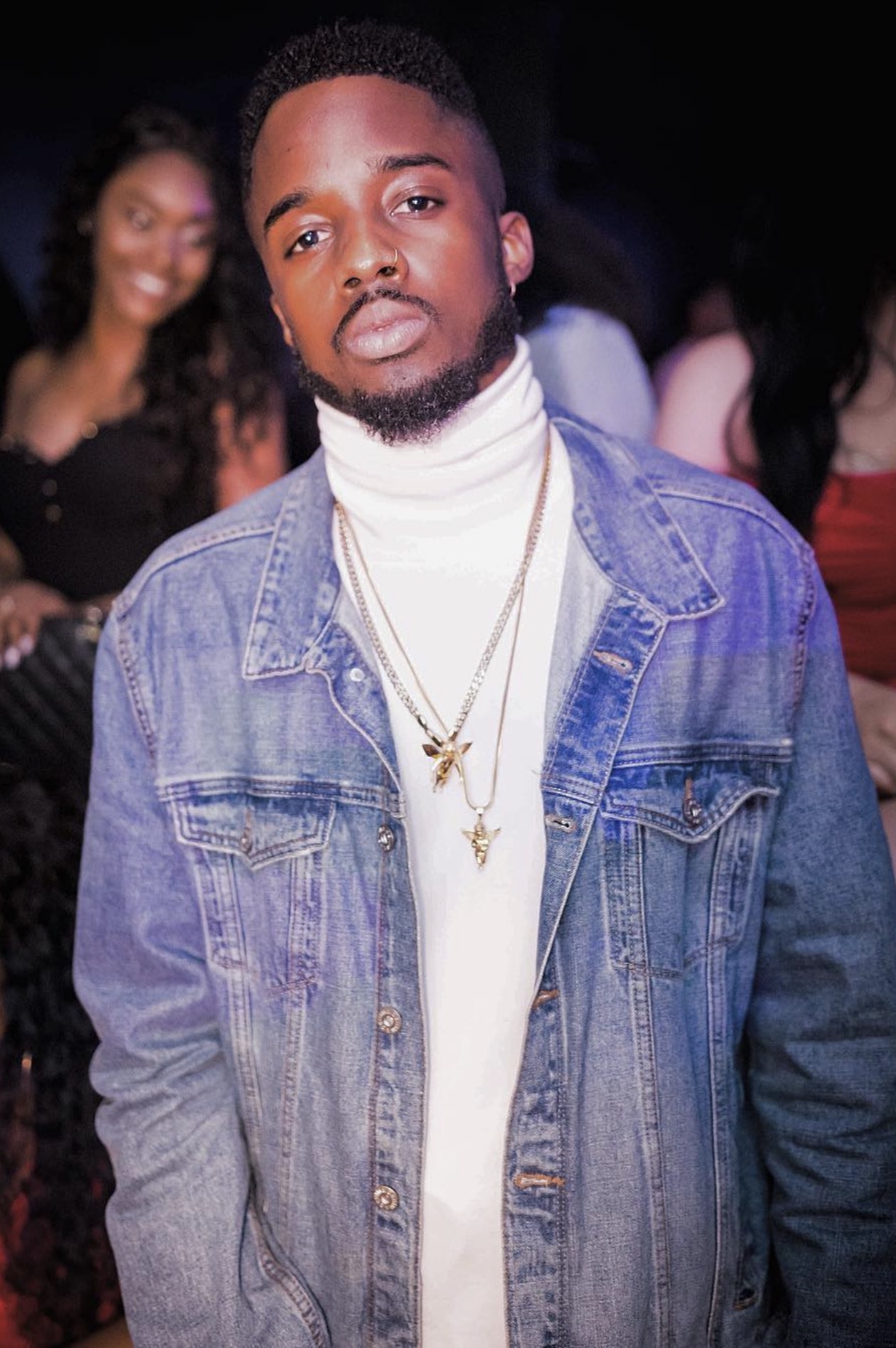 Young man with a beard wearing a denim jacket and white turtleneck, standing indoors with people in the background.