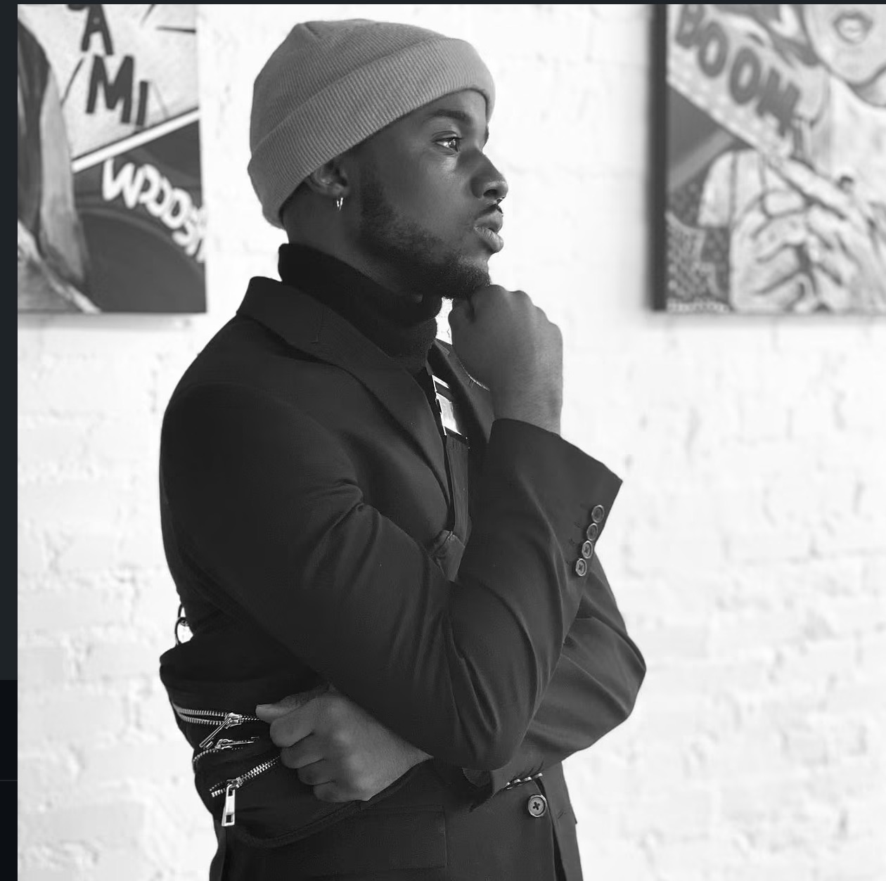 Young man in a beanie and jacket, standing in profile with hand on chin, in front of a white brick wall with artwork.