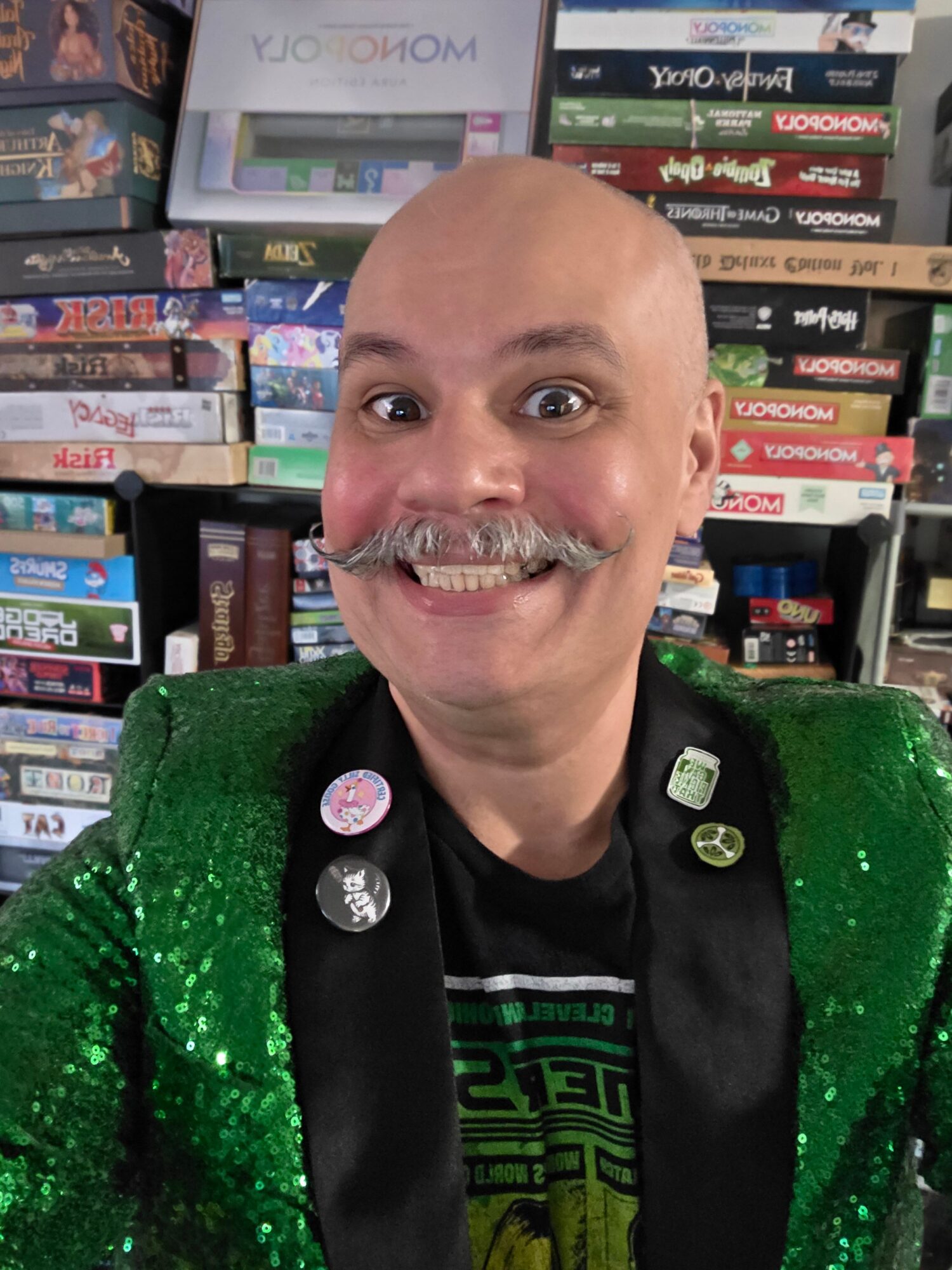 Bald man with a mustache smiling, wearing a green sequined jacket and black shirt, in front of a bookshelf