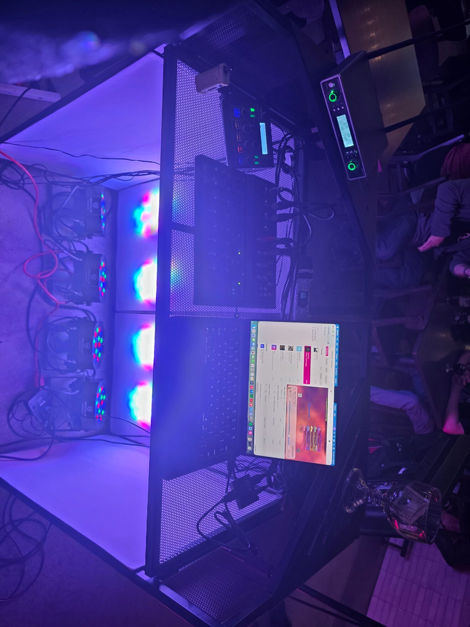 A DJ setup with a laptop, mixer, and equipment on a table illuminated by colorful lights, in a dimly lit room.