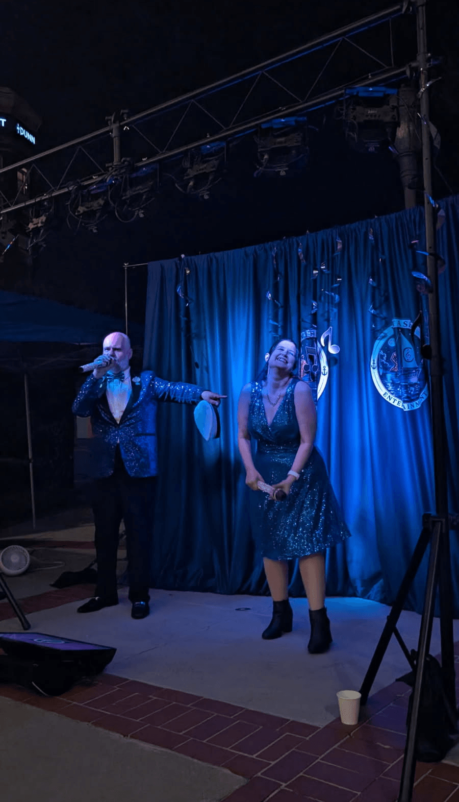 Two performers on stage with blue curtains, one singing into a microphone, the other standing with hands clasped.