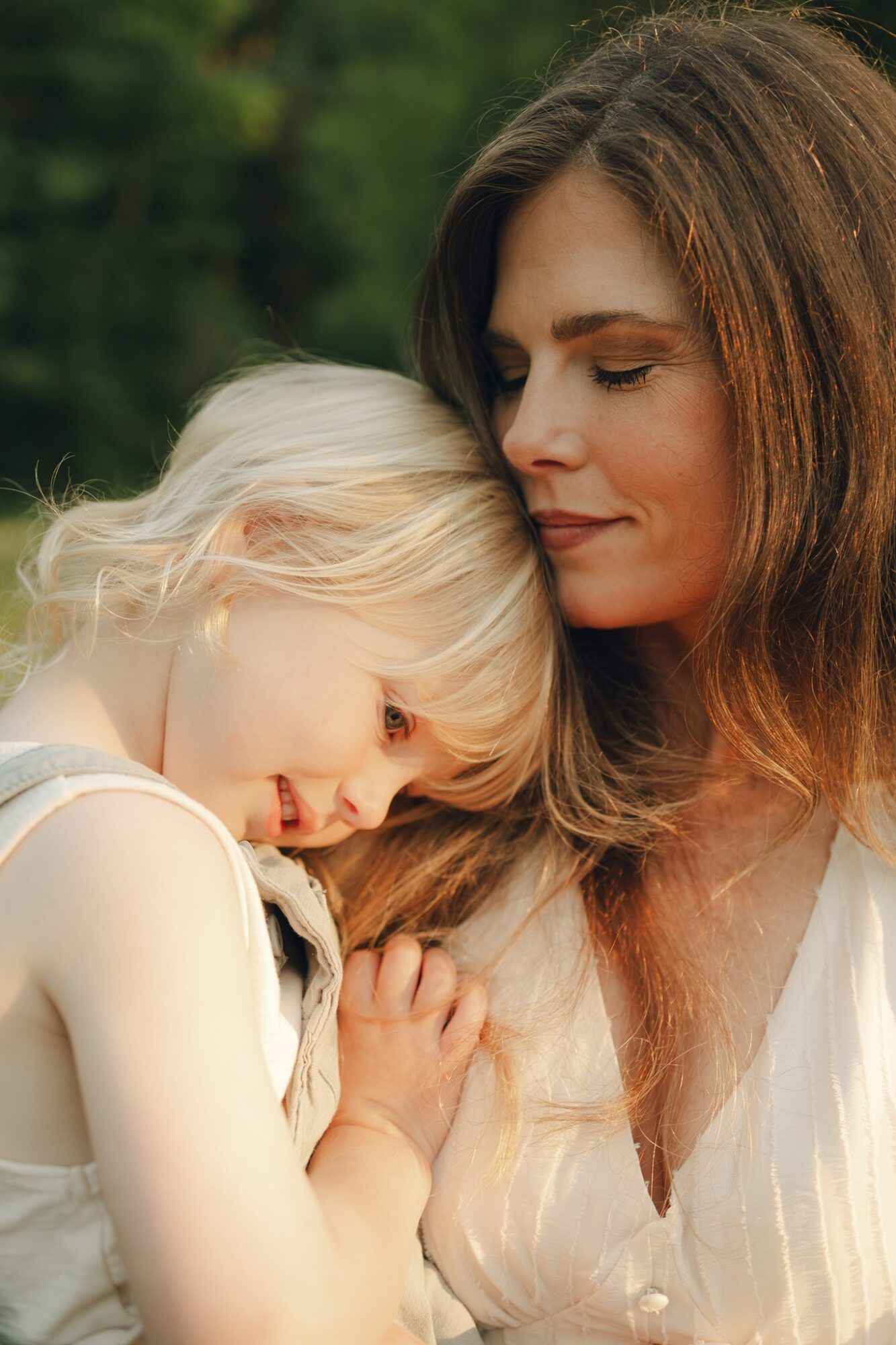 A woman and a young girl with blonde hair are close together outdoors, with the girl resting her head on the woman's shoulder.