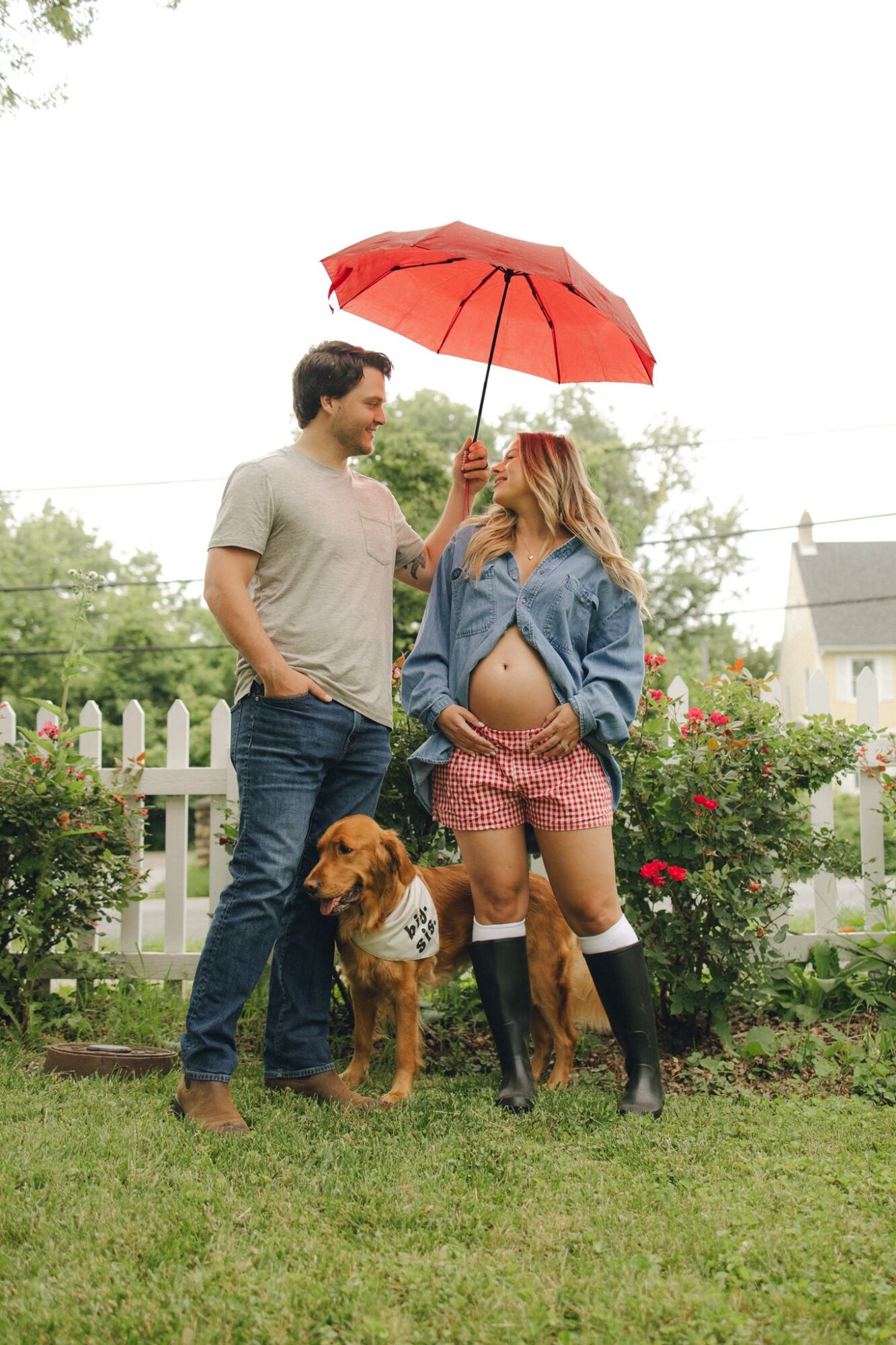 A pregnant woman holding a red umbrella, a man, and a dog in a garden with a white fence and flowers.