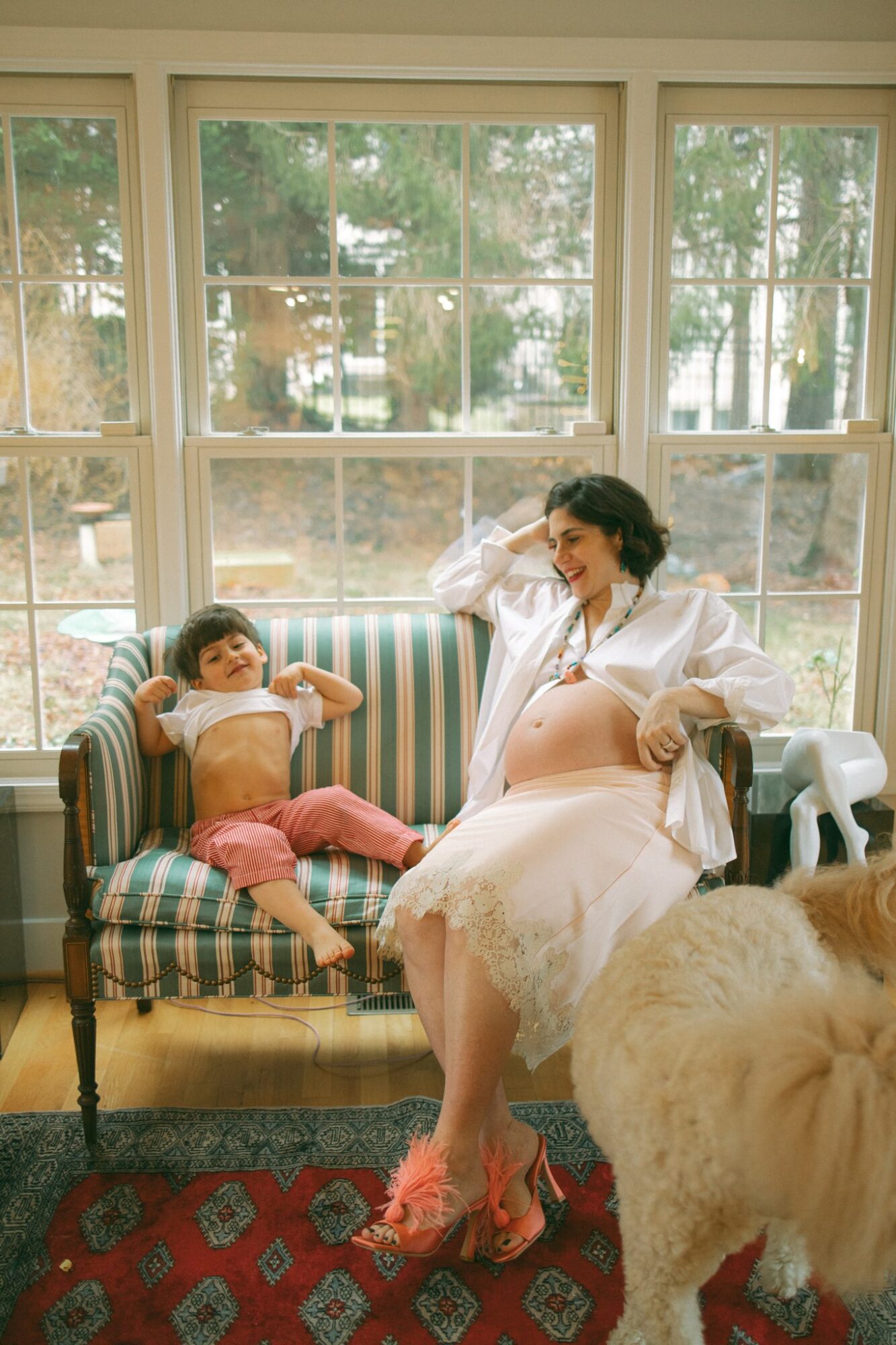 Pregnant woman and young girl sitting on a striped sofa by large windows, with a dog nearby, in a bright room.