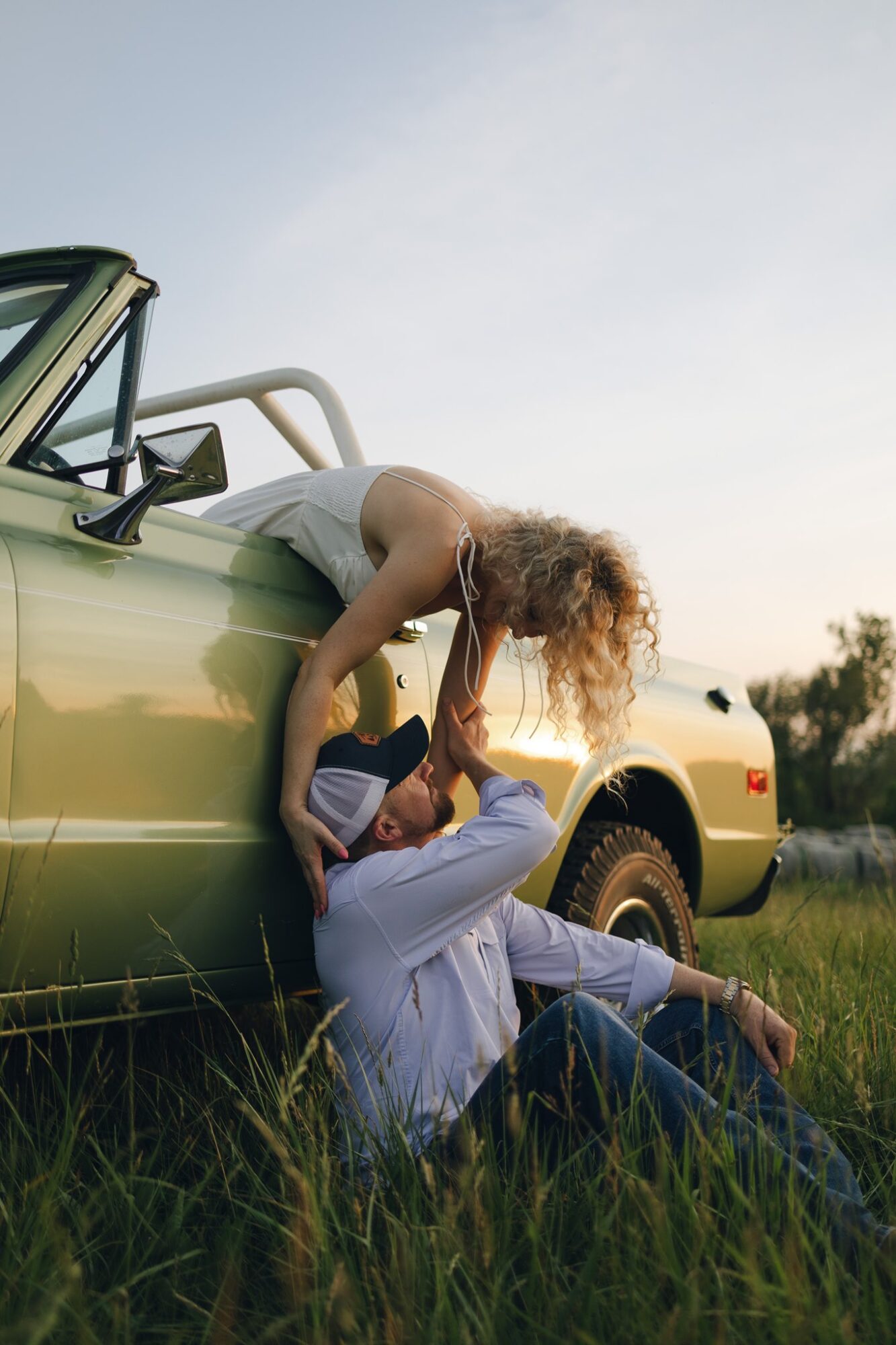 Two people near a green car in a grassy field, one lying on the ground and the other leaning over, with a clear sky.