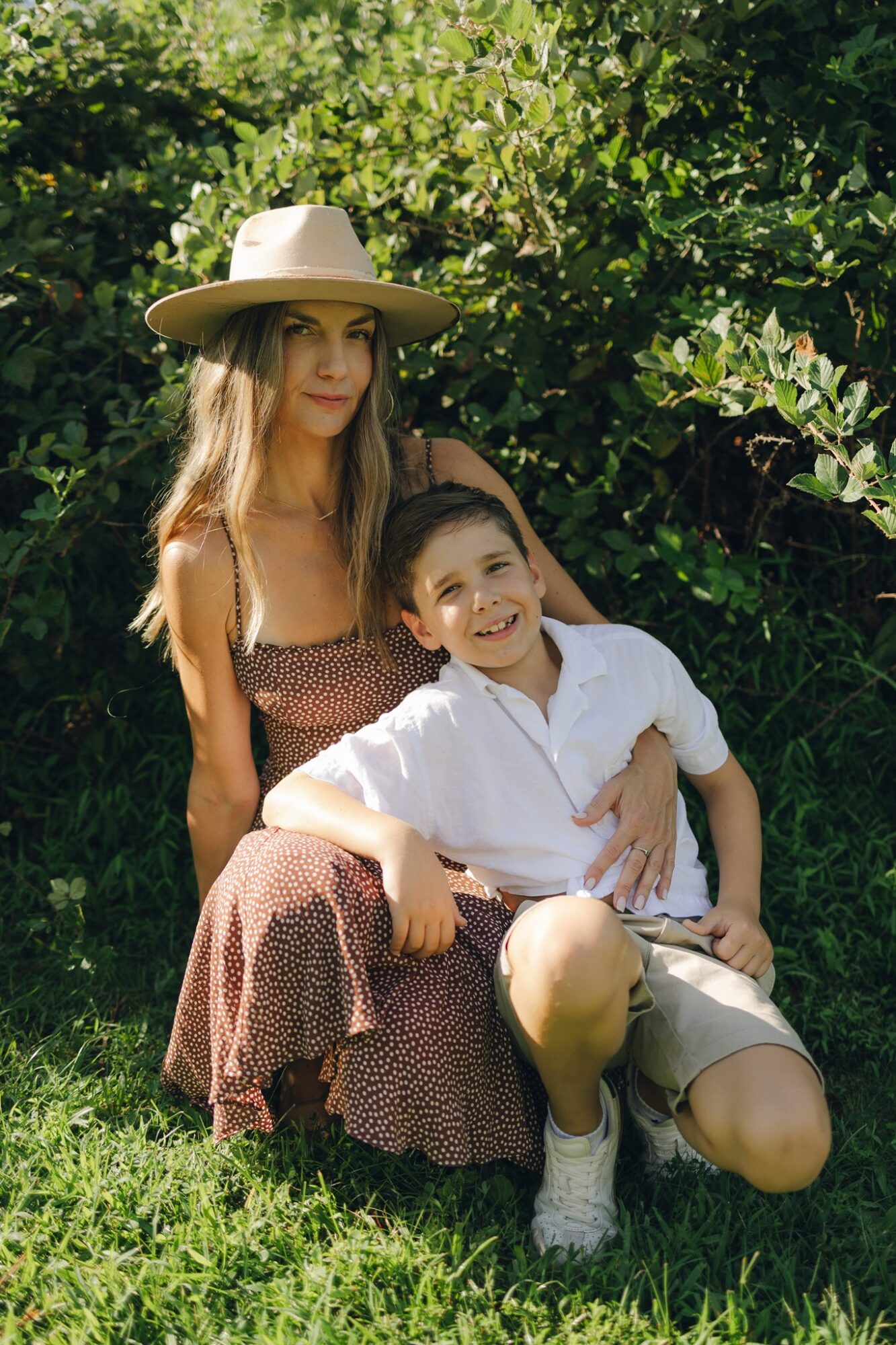 Woman and child sitting outdoors on grass, surrounded by greenery, woman wearing a wide-brimmed hat and dress, child in white shirt and shorts.