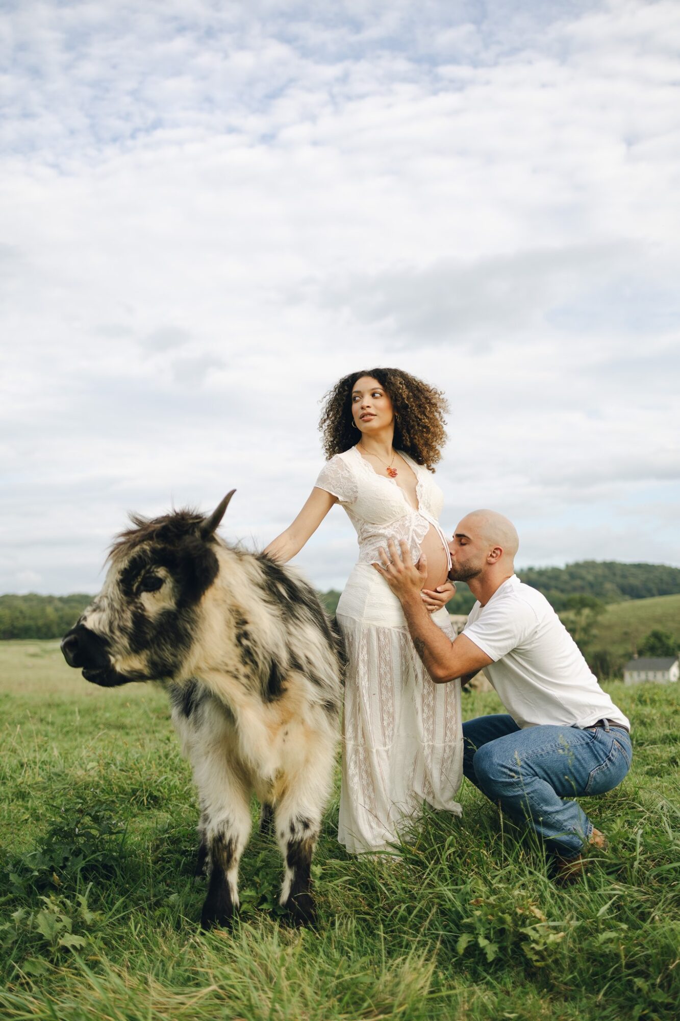 A woman with curly hair in a white dress stands outdoors with a man kneeling beside her, holding her hand, and a small pony nearby.