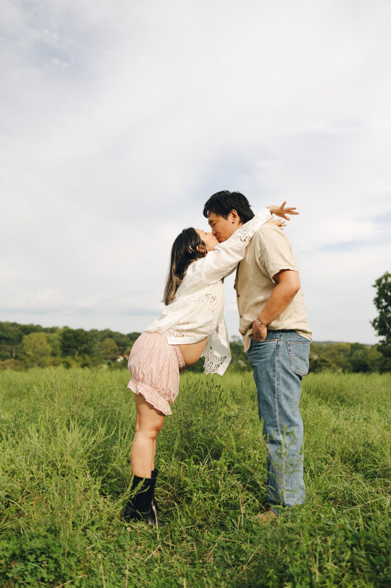A couple stands close in a grassy field, leaning in for a kiss under a cloudy sky.