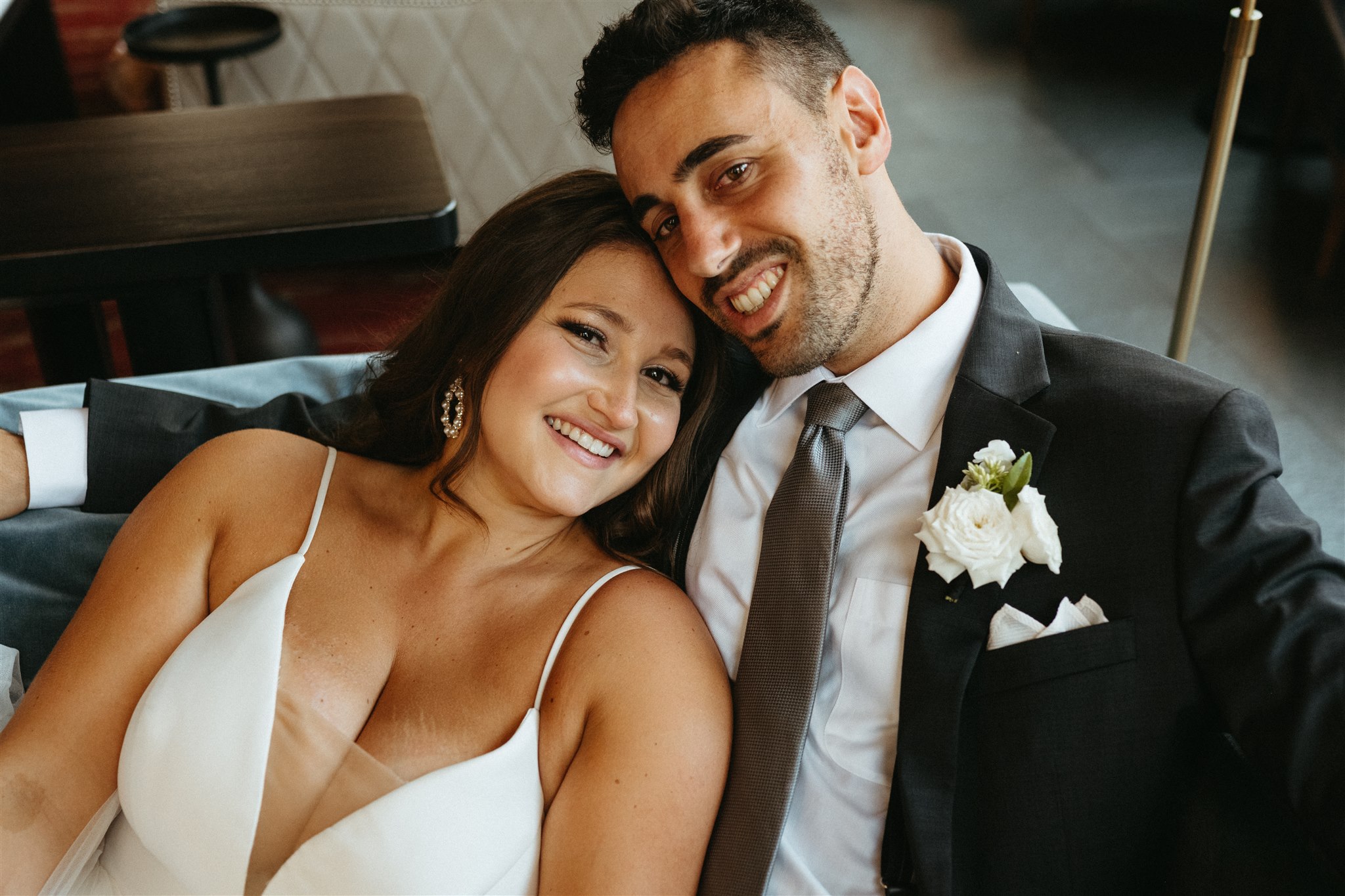 A smiling couple in wedding attire, close together indoors, with the woman in a white dress and the man in a suit.