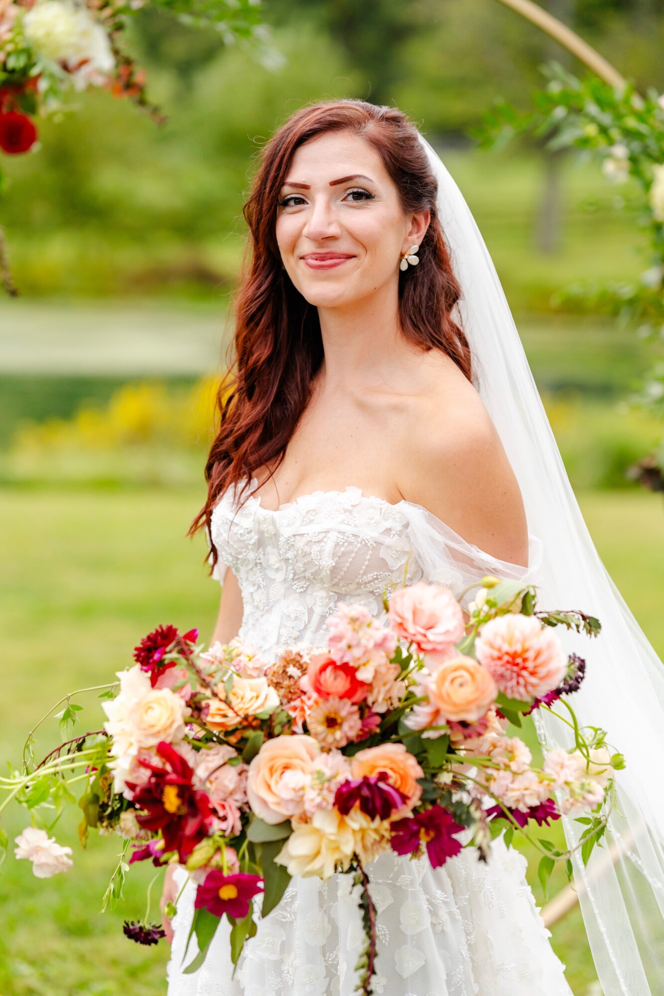 Bride in wedding dress holding a large bouquet outdoors, smiling, with greenery in background.