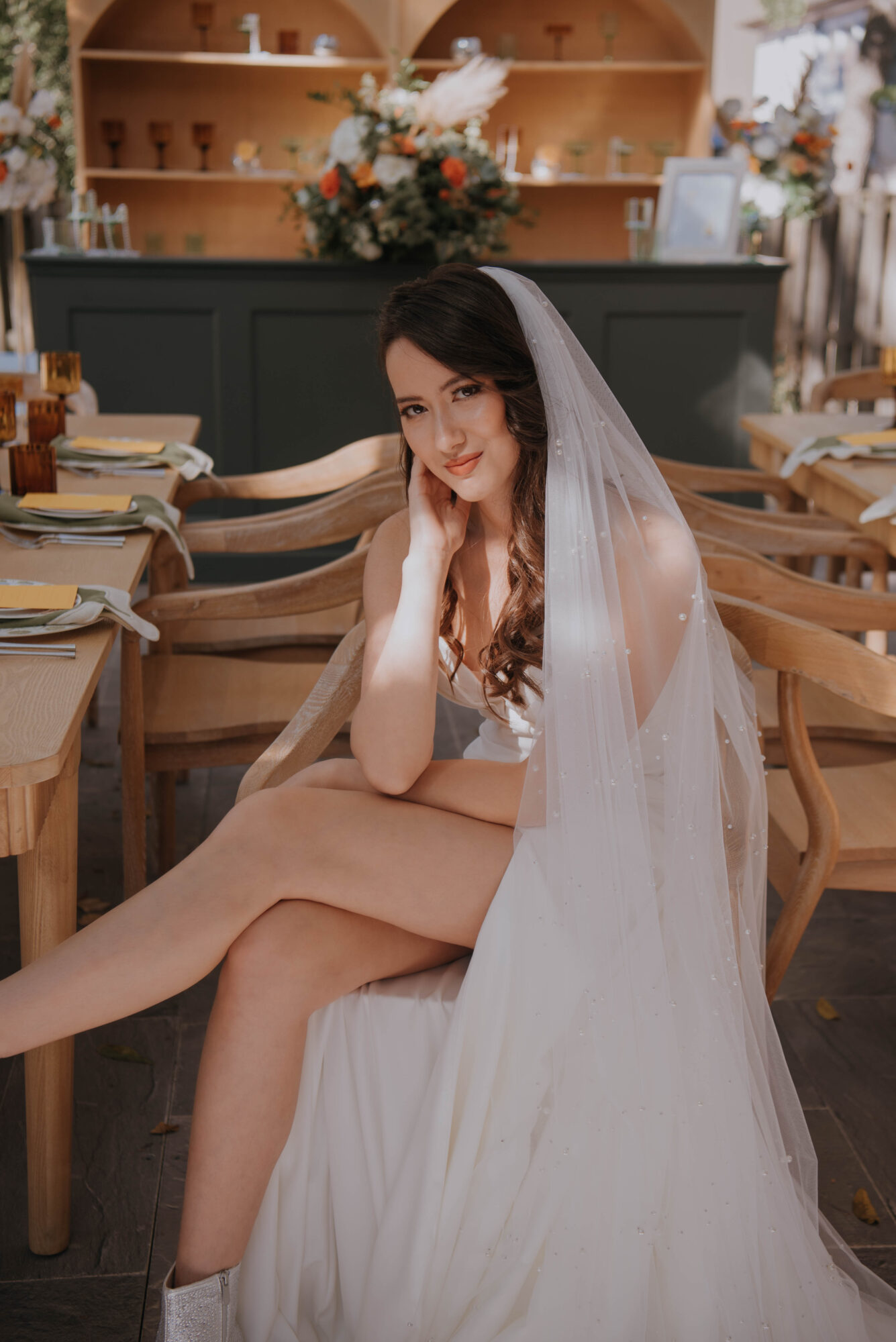 Young woman in wedding dress sitting at table with veil, smiling, in a decorated indoor setting.