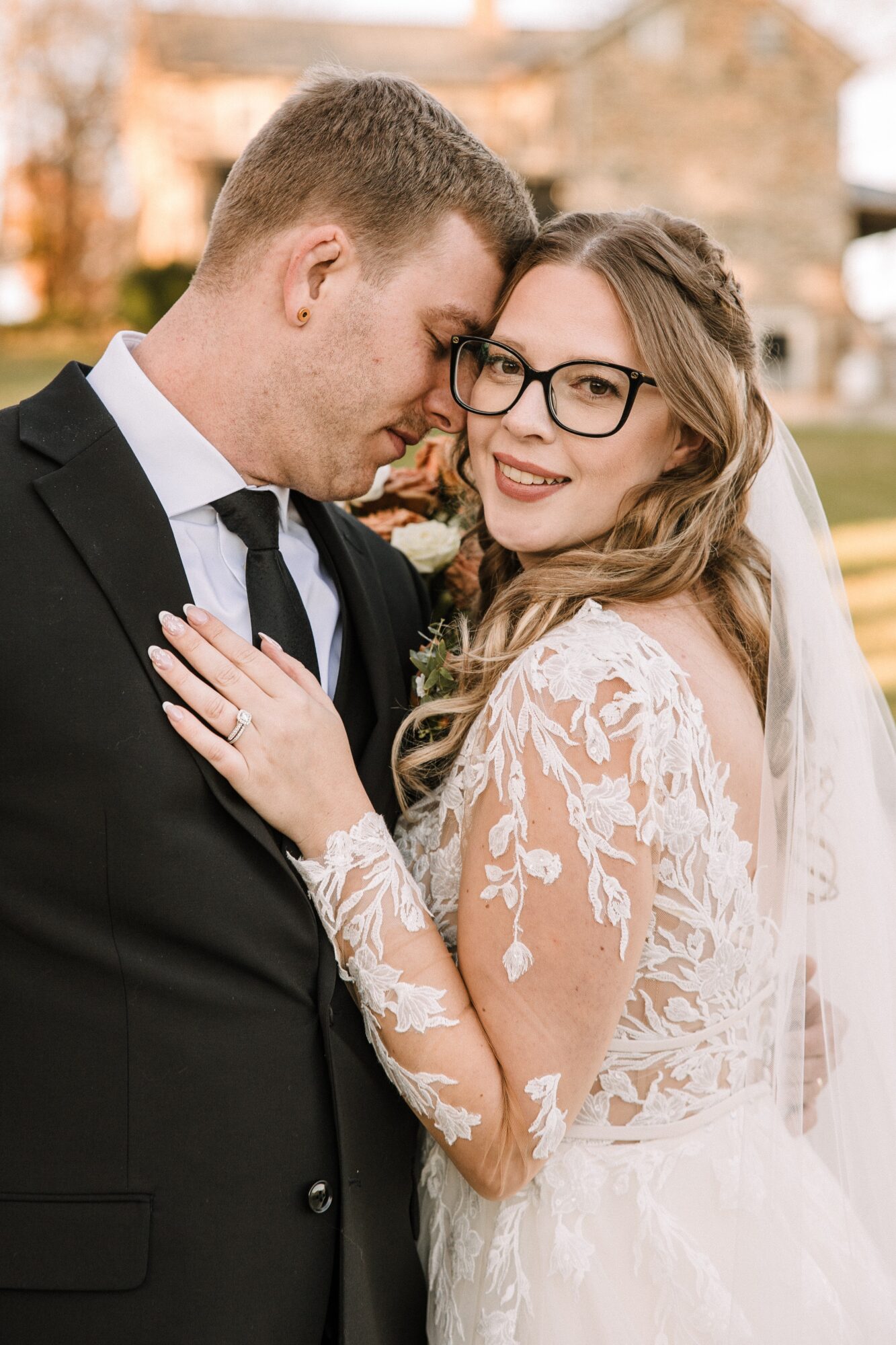 A bride and groom close together, smiling, outdoors with blurred background, woman wearing glasses and lace wedding dress.