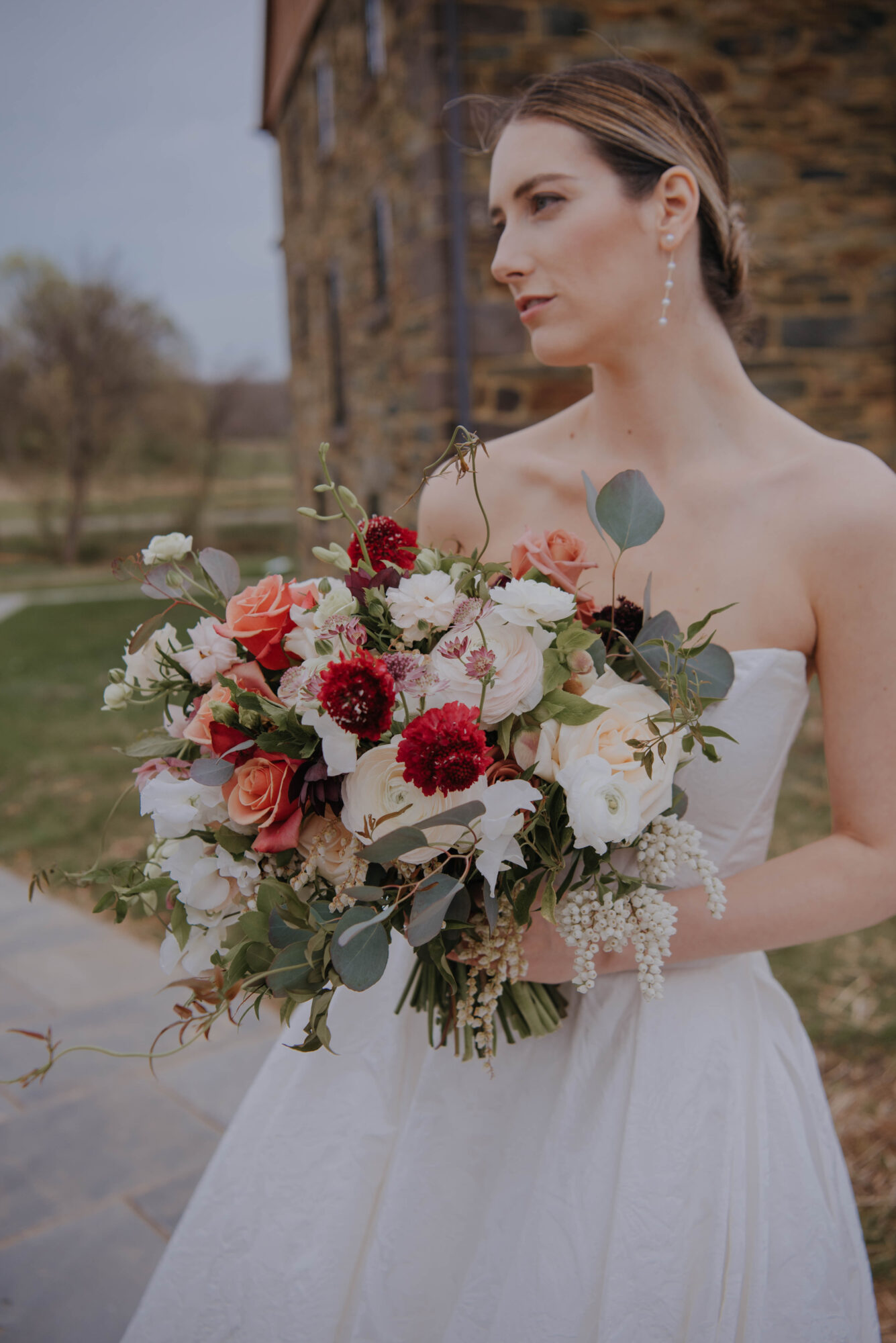 Bride holding a large bouquet of flowers outdoors, with a stone building in the background.
