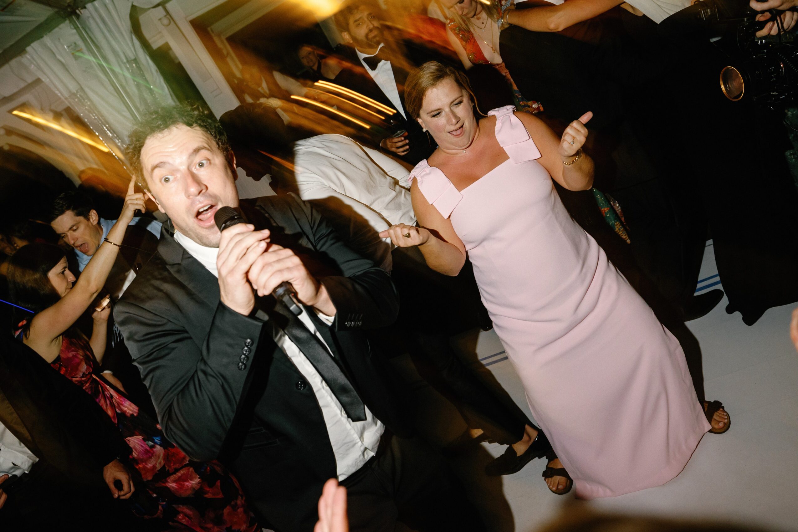 Man singing into a microphone and woman dancing at a party with people in the background.