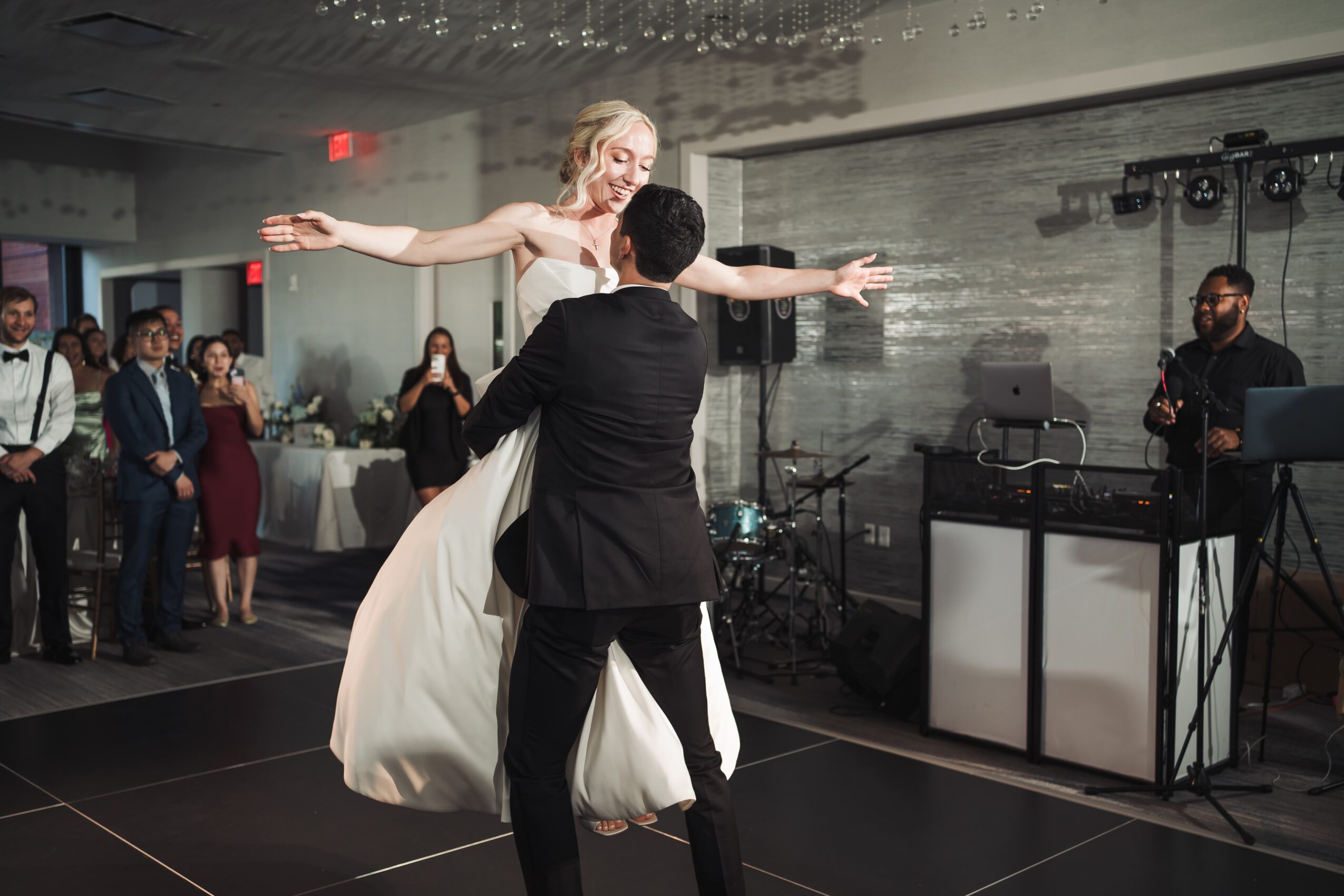 A bride and groom dance together at a wedding reception, with guests watching in the background and a DJ on the right.