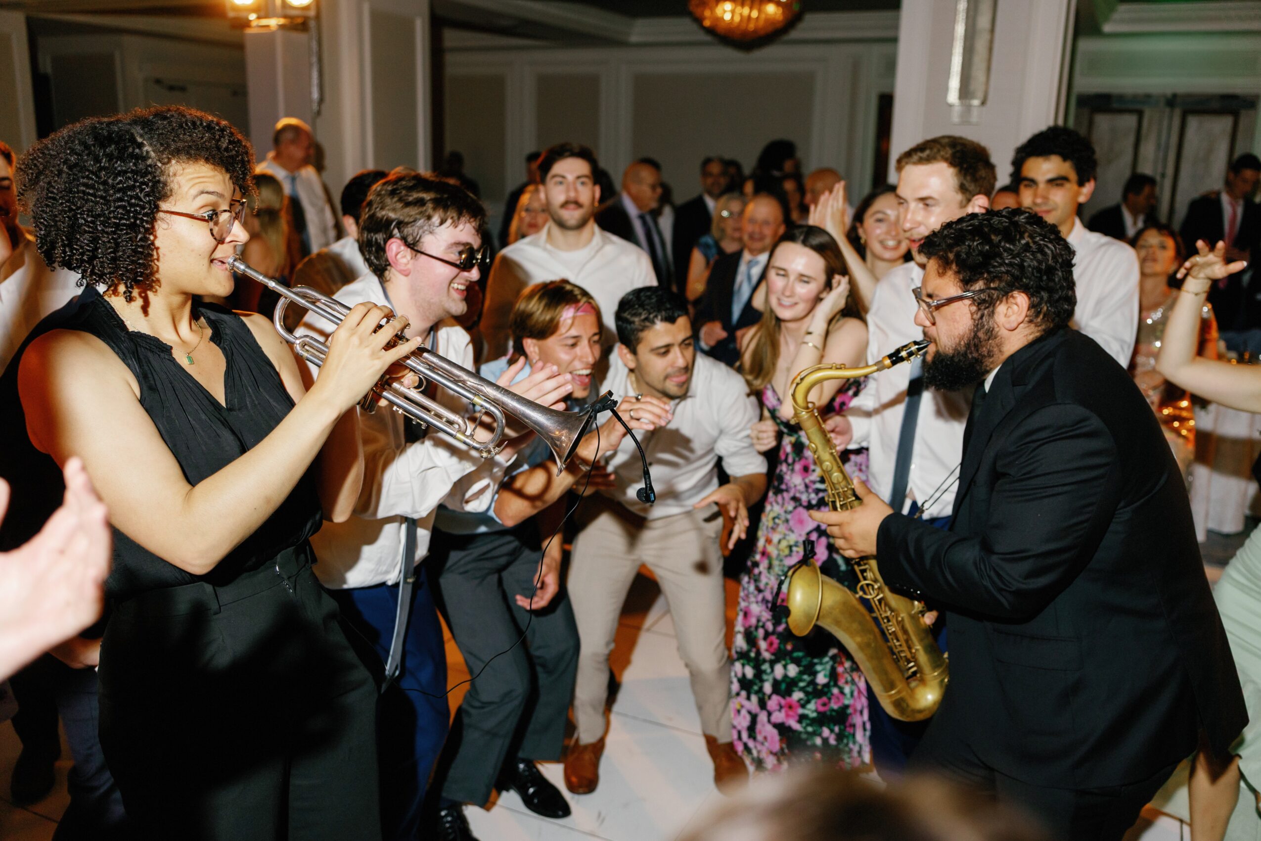 Group of people dancing and enjoying live music with a woman playing trumpet and a man playing saxophone, in an indoor event.