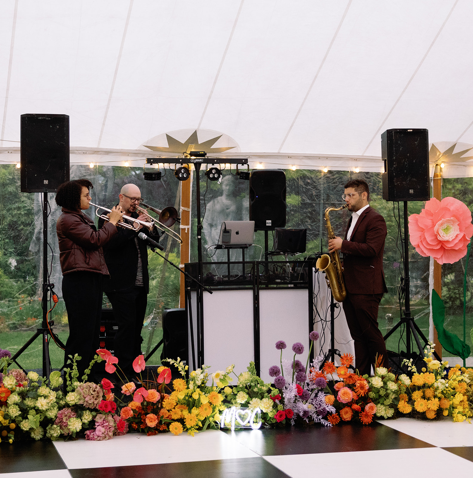 Three musicians perform on stage with flowers, speakers, and decorations, under a white canopy. One plays trumpet, another plays saxophone, and a third is at a DJ setup.