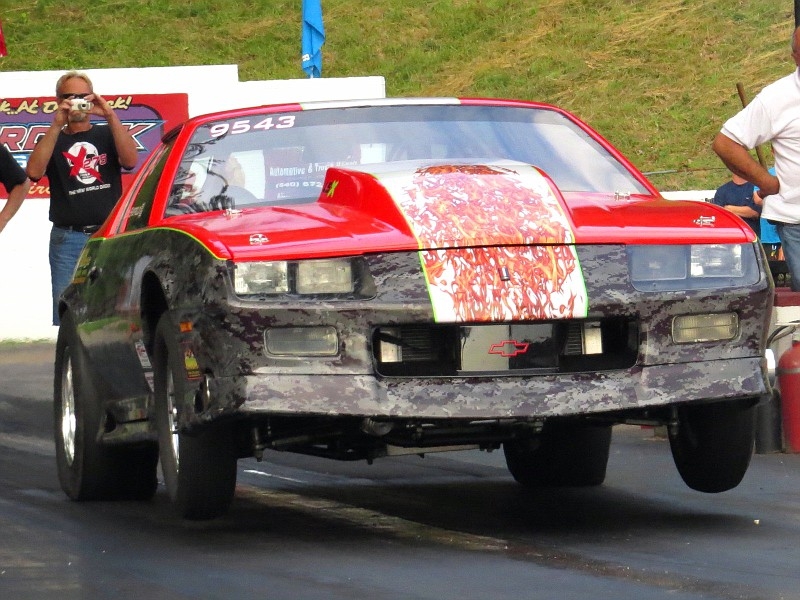 Red race car with damage and a large white stripe, on a track with spectators nearby.