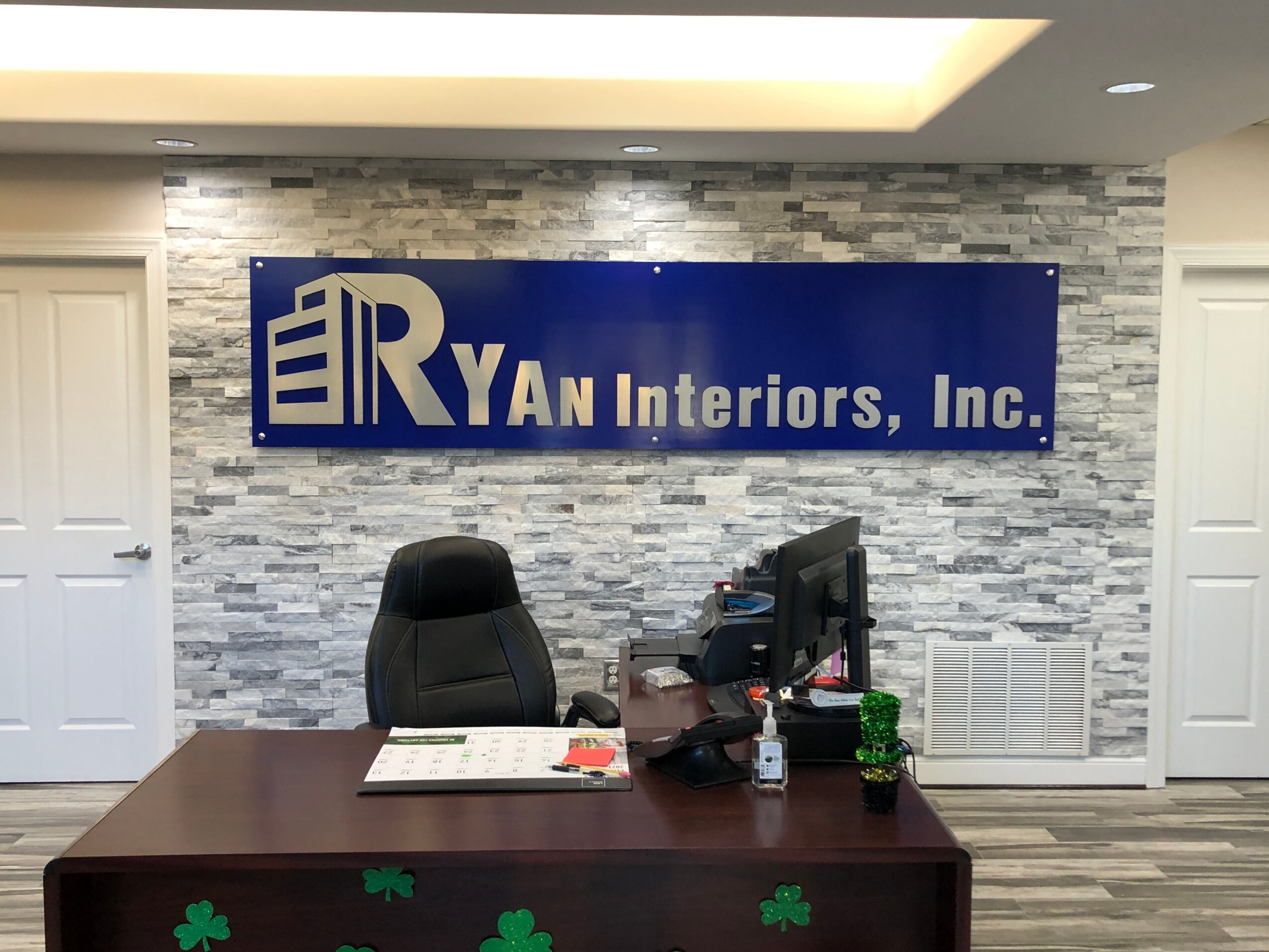 Office reception area with a desk, chair, computer, and a blue sign reading 'RYAN Interiors, Inc.' on a textured wall.