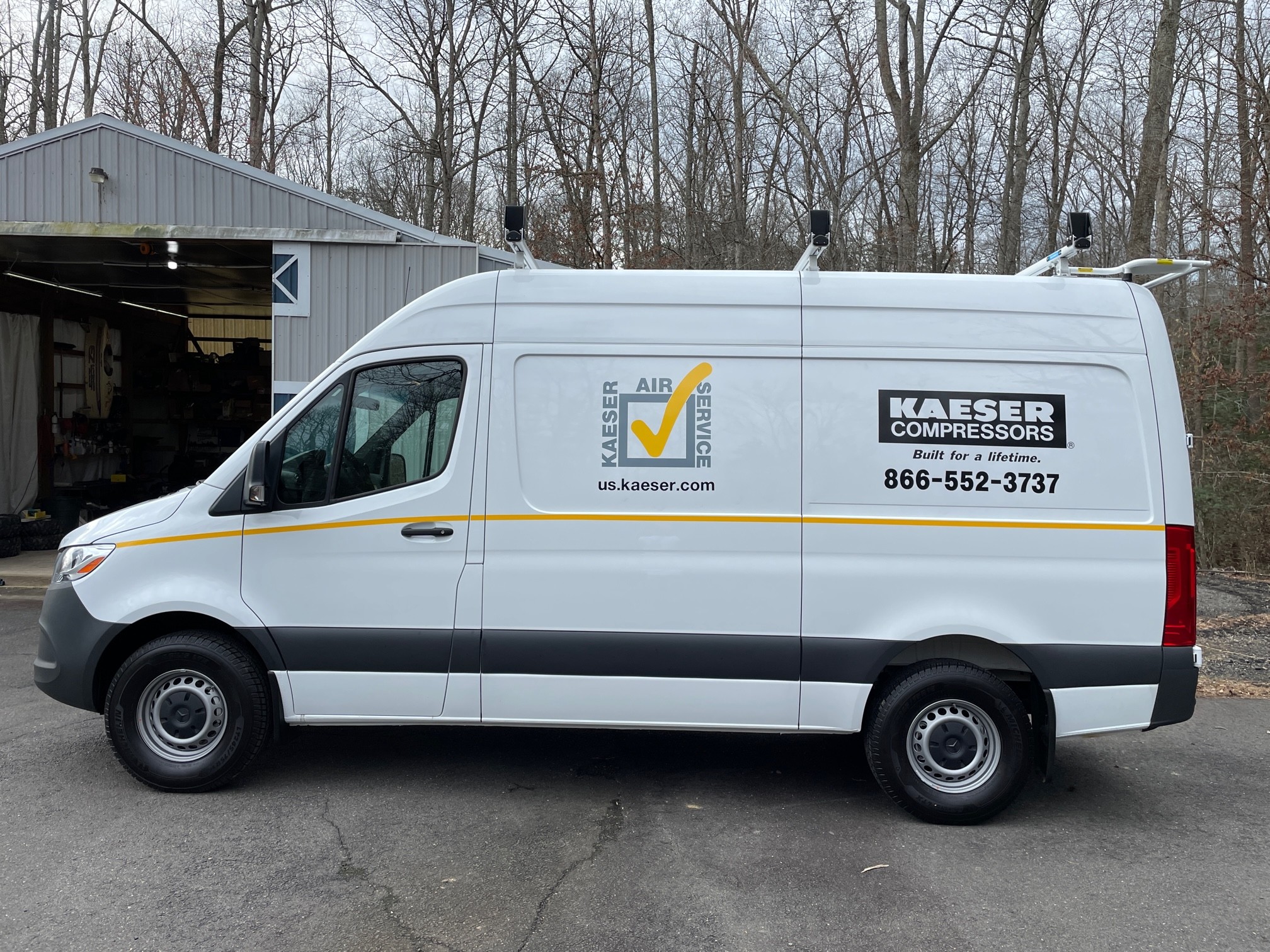 White delivery van with company logo and contact information parked outside, trees and a building in background.