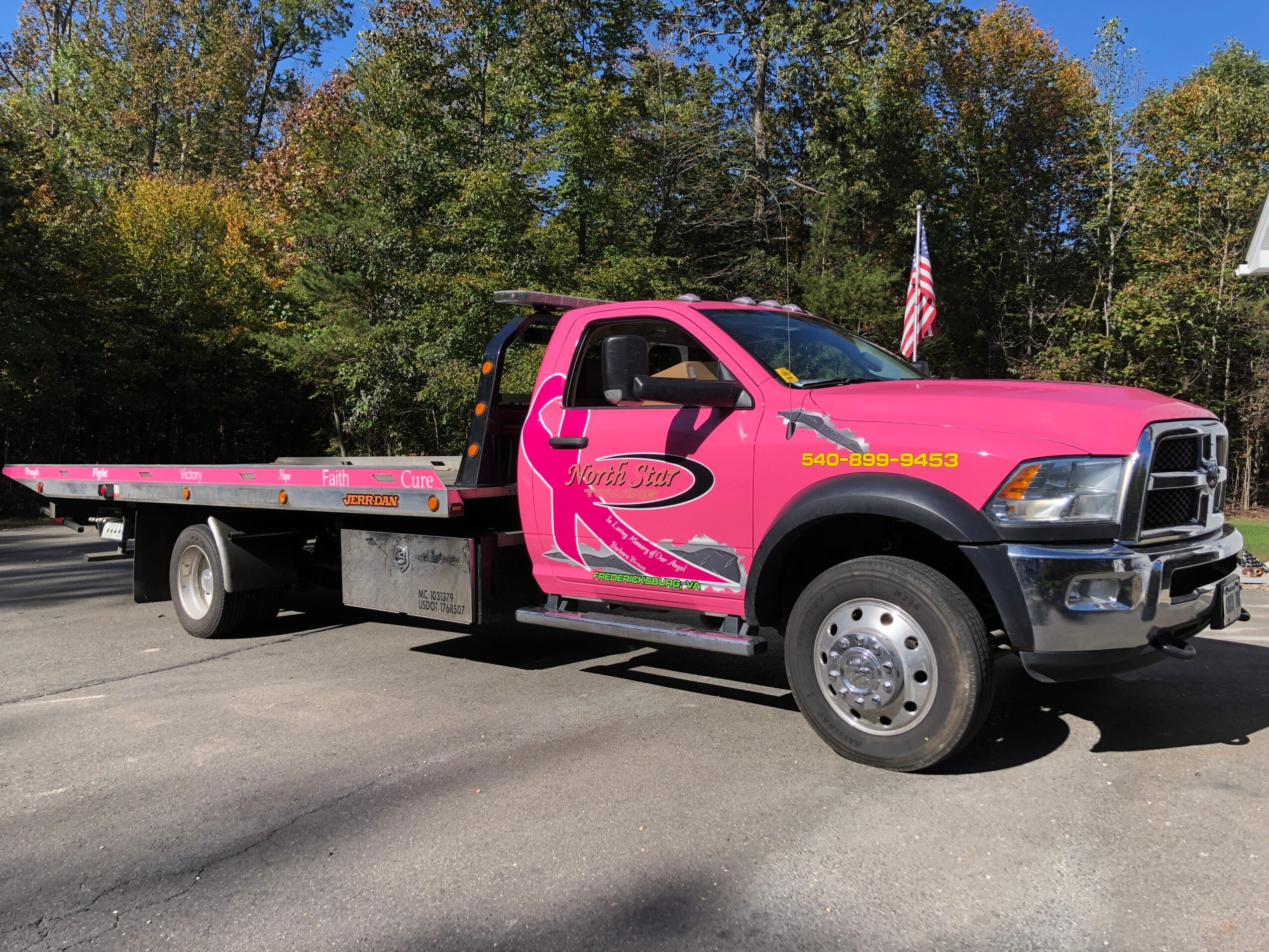 Pink tow truck parked outdoors with trees in the background and an American flag nearby.