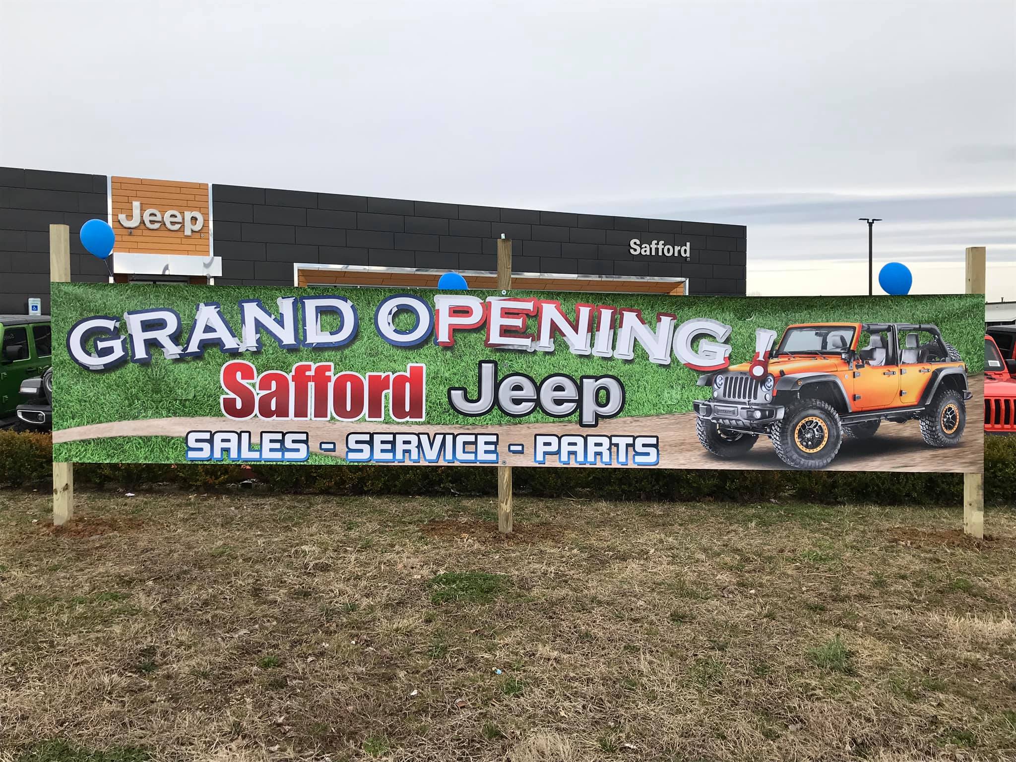Grand opening banner for Safford Jeep with a vehicle image, on grass, in front of a dealership building, overcast sky.
