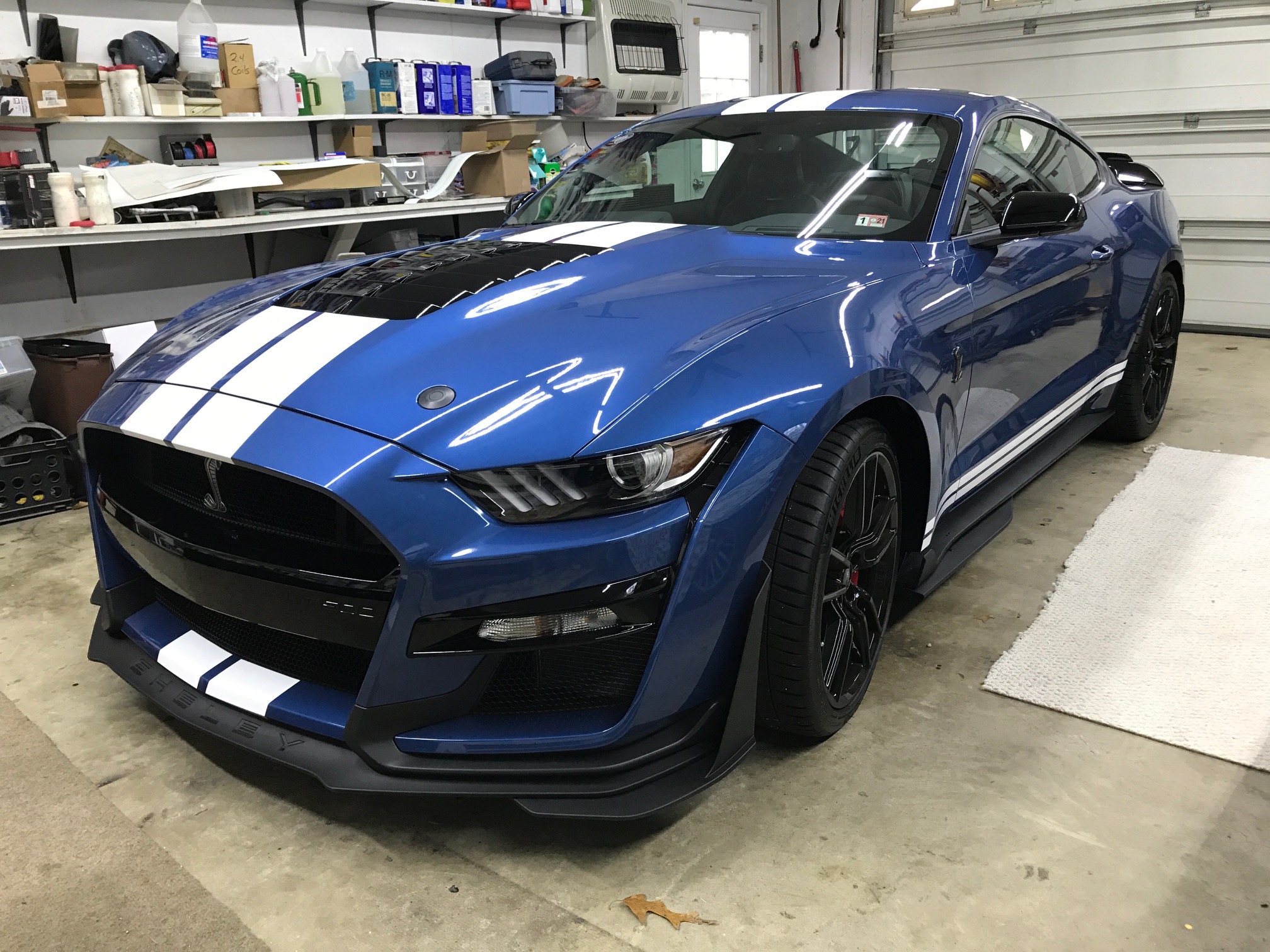 Blue sports car with white racing stripes parked inside a garage, surrounded by shelves and tools.
