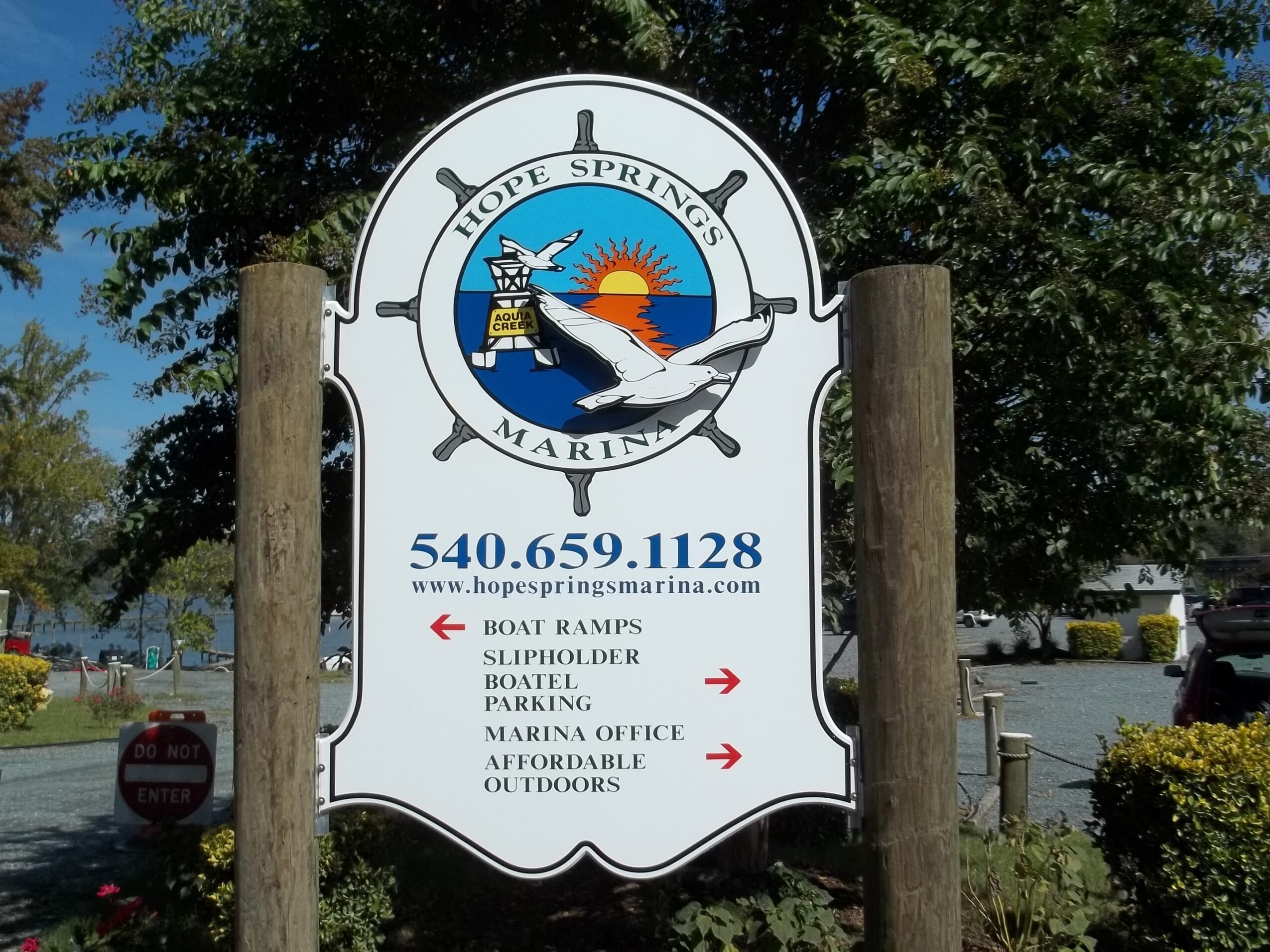 Sign for Hope Springs Marina with a dolphin, boat, and sunset, listing contact info and amenities, outdoors with trees and cars.
