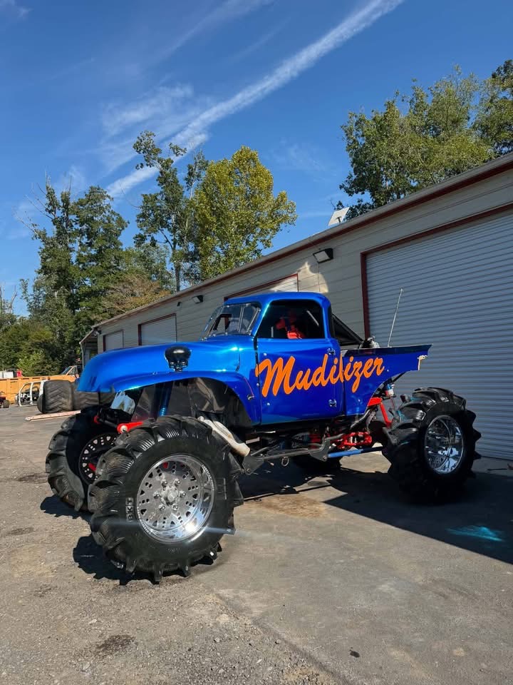 Blue monster truck with large tires and orange 'Mudlizer' logo on side, parked outside a building under a blue sky.