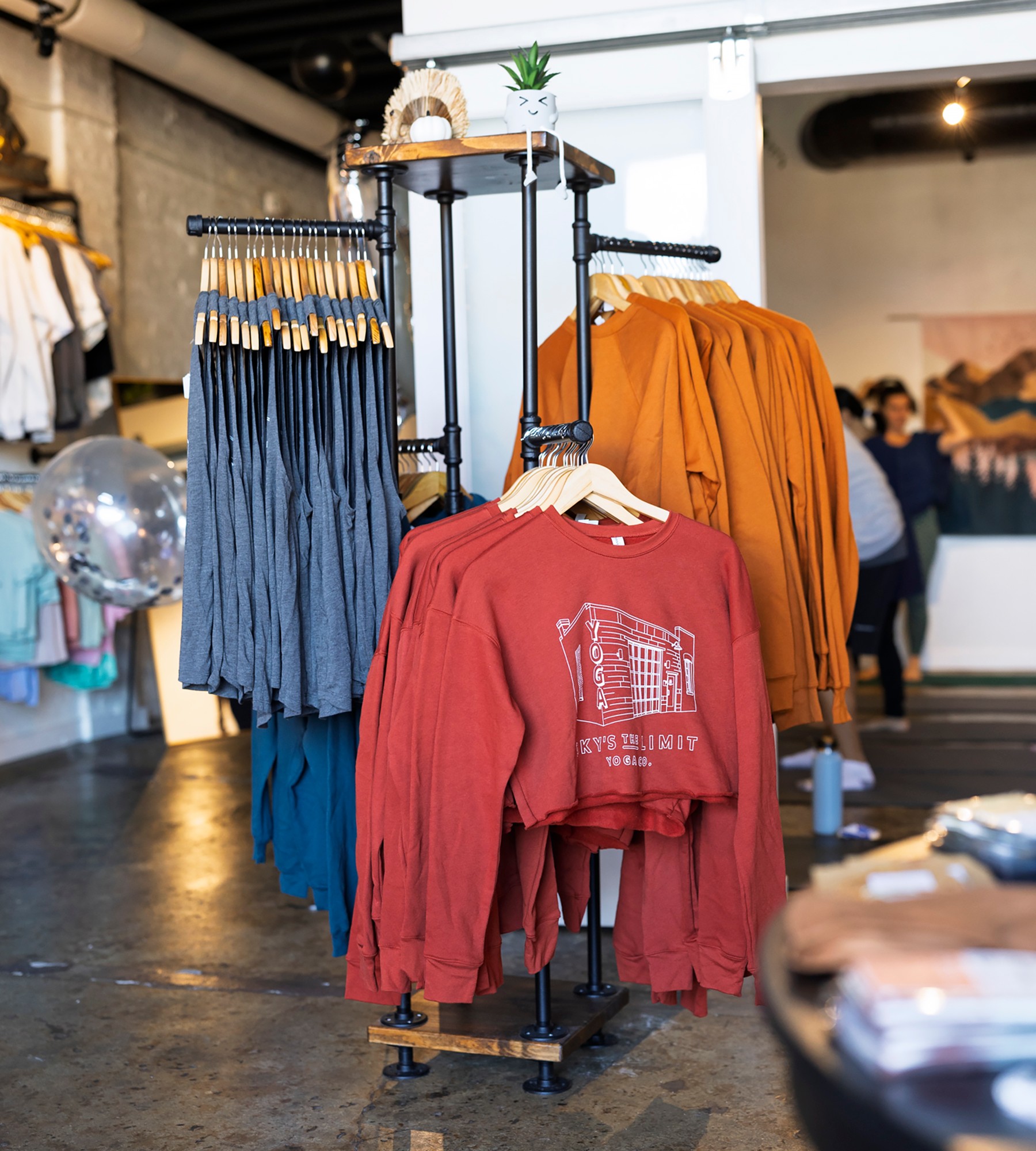 Clothing rack with red, orange, and blue garments in a retail store, with people in the background. 20 words.