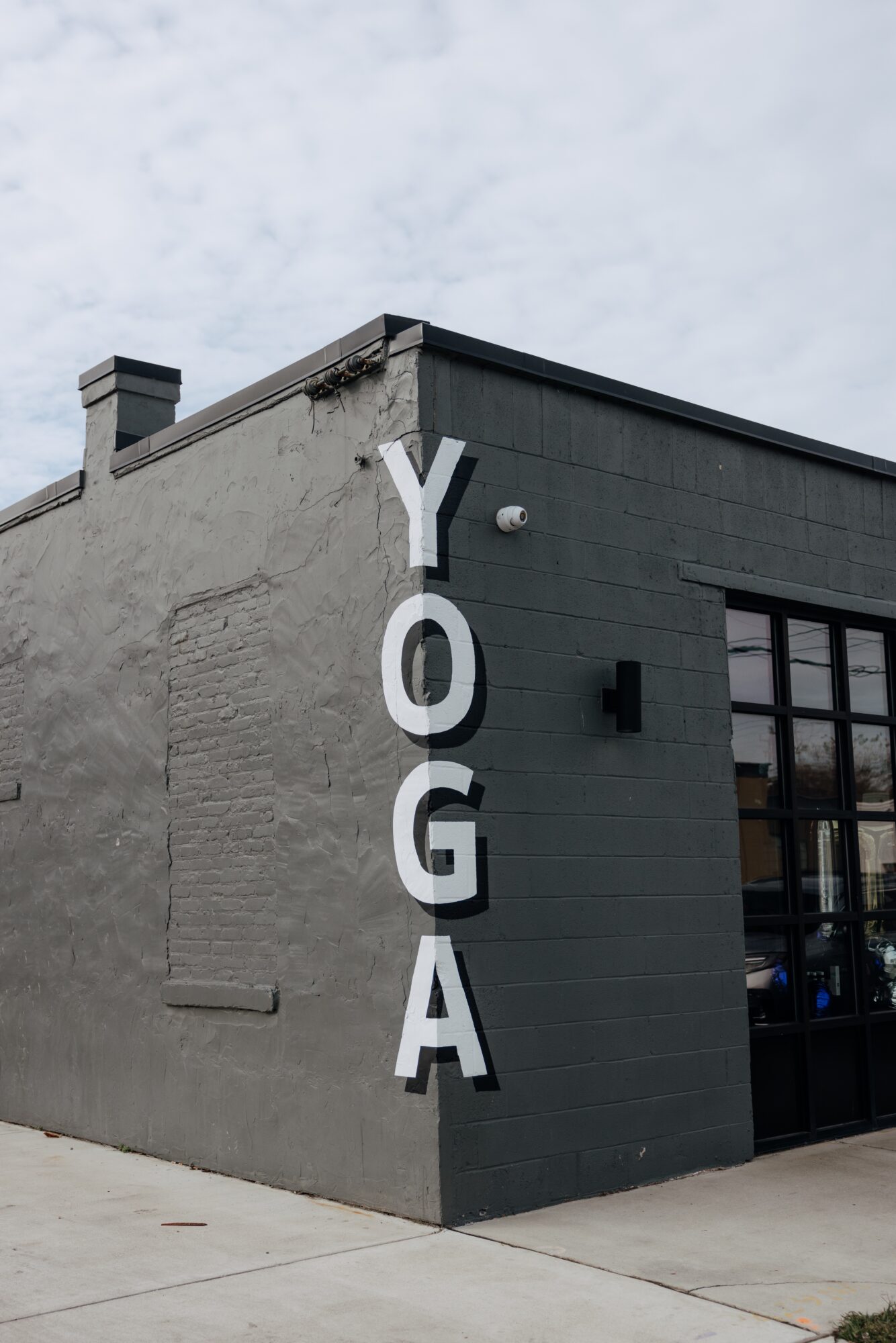 Corner of a building with large vertical white letters spelling 'YOGA' on a gray wall, under a cloudy sky.