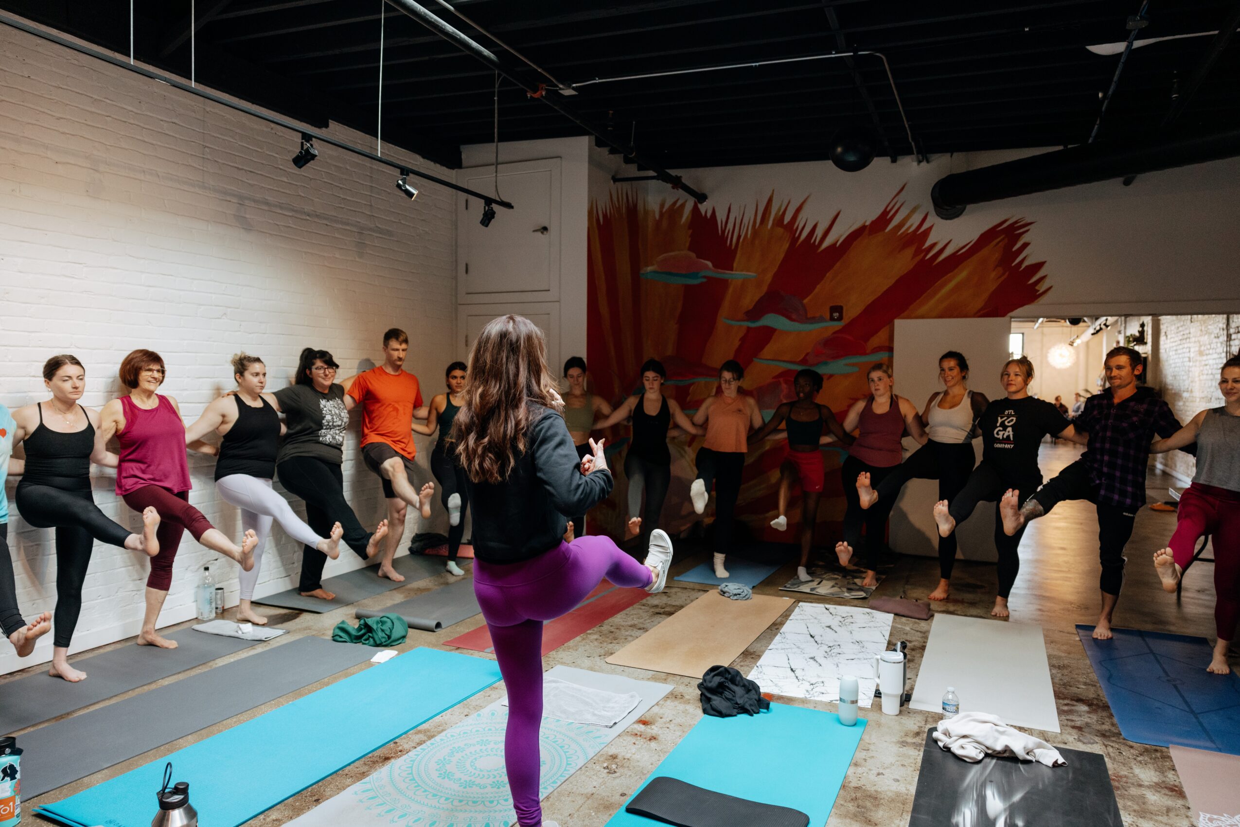 Group of people participating in a fitness class, standing on mats with one leg raised, in a studio with colorful wall art.