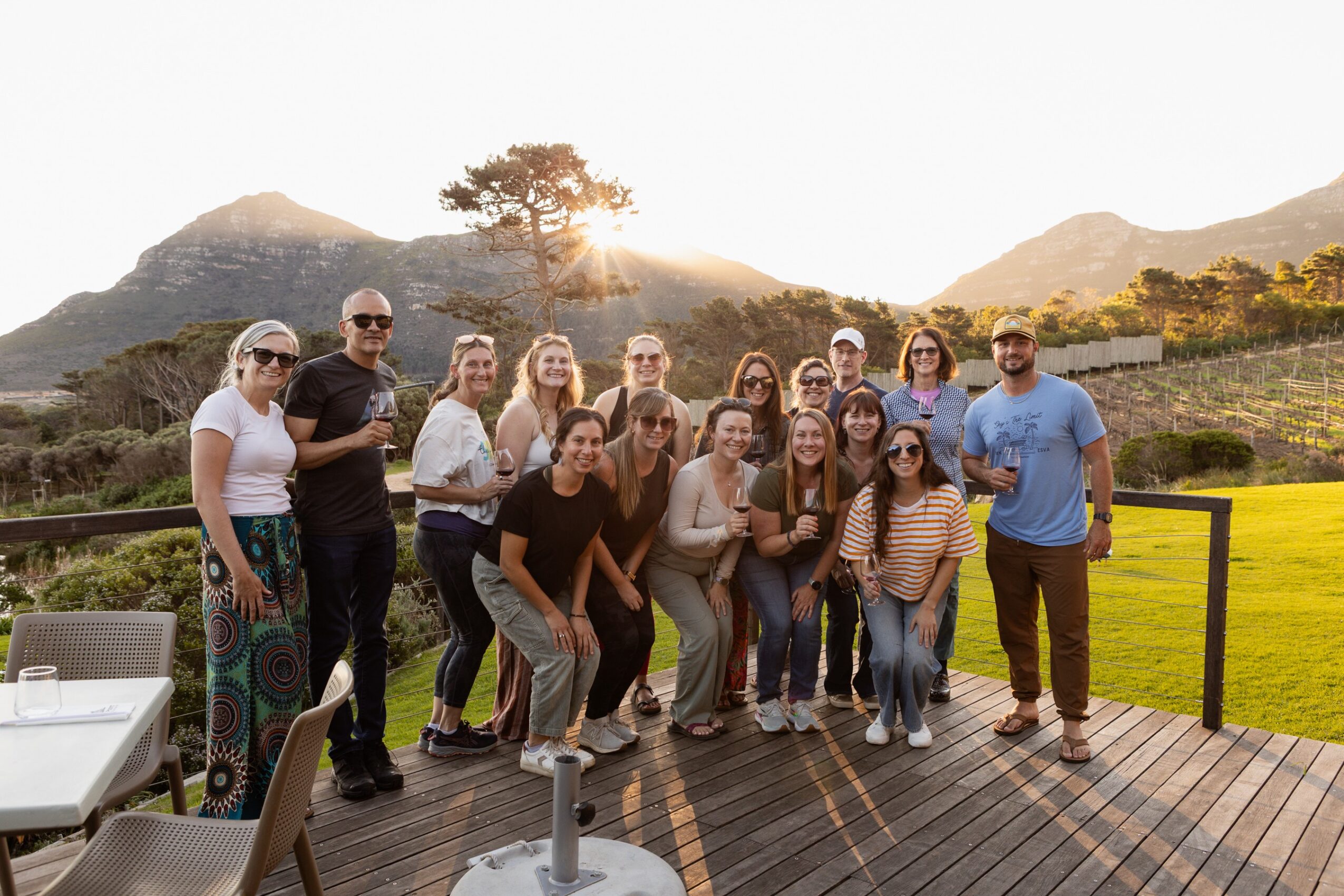 Group of people outdoors on a wooden deck with mountains and a tree in the background, some smiling and posing for the photo.